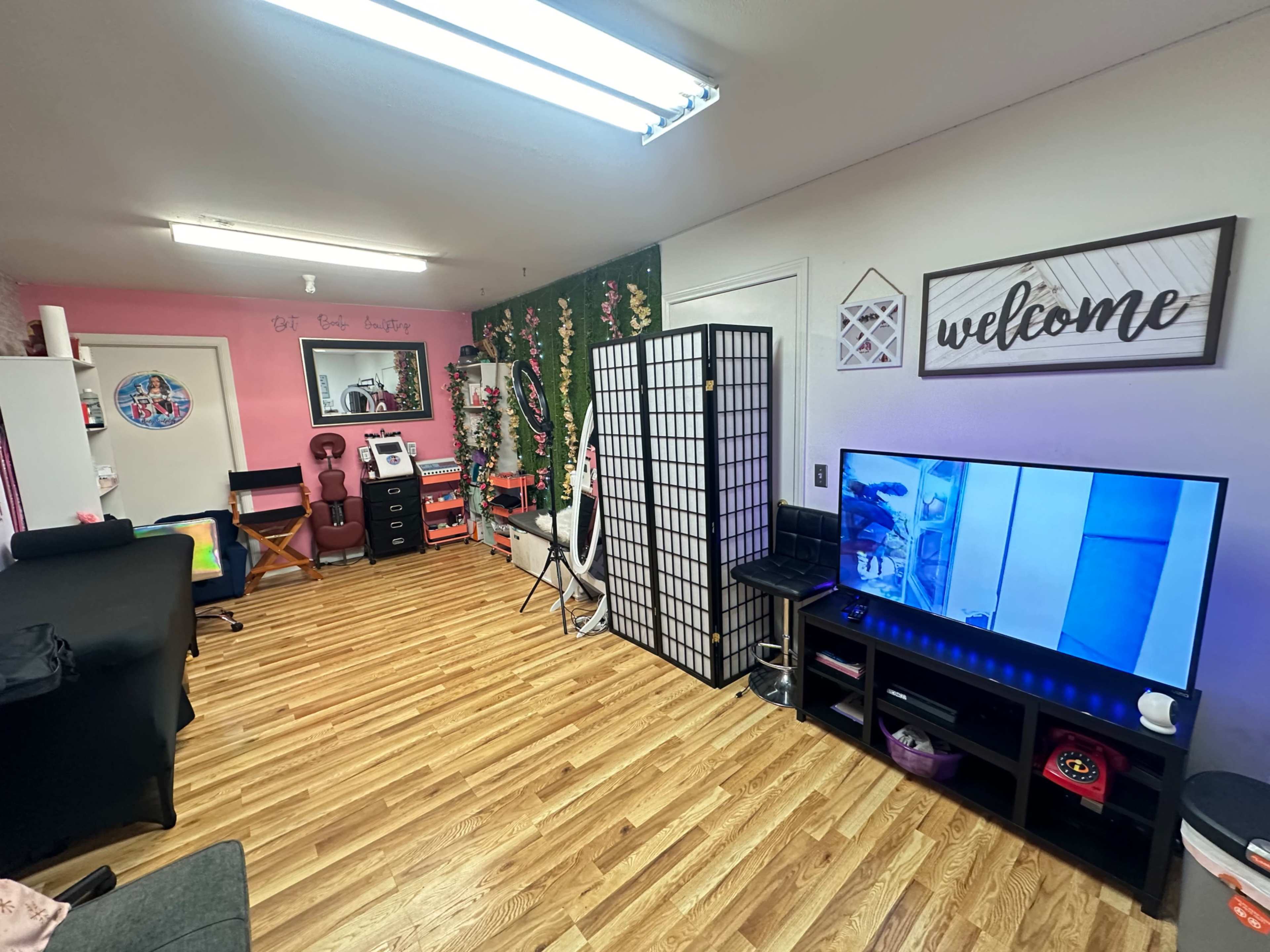 The image depicts a brightly lit room with wooden flooring, featuring a television, a decorative screen, and various furniture items along the walls.