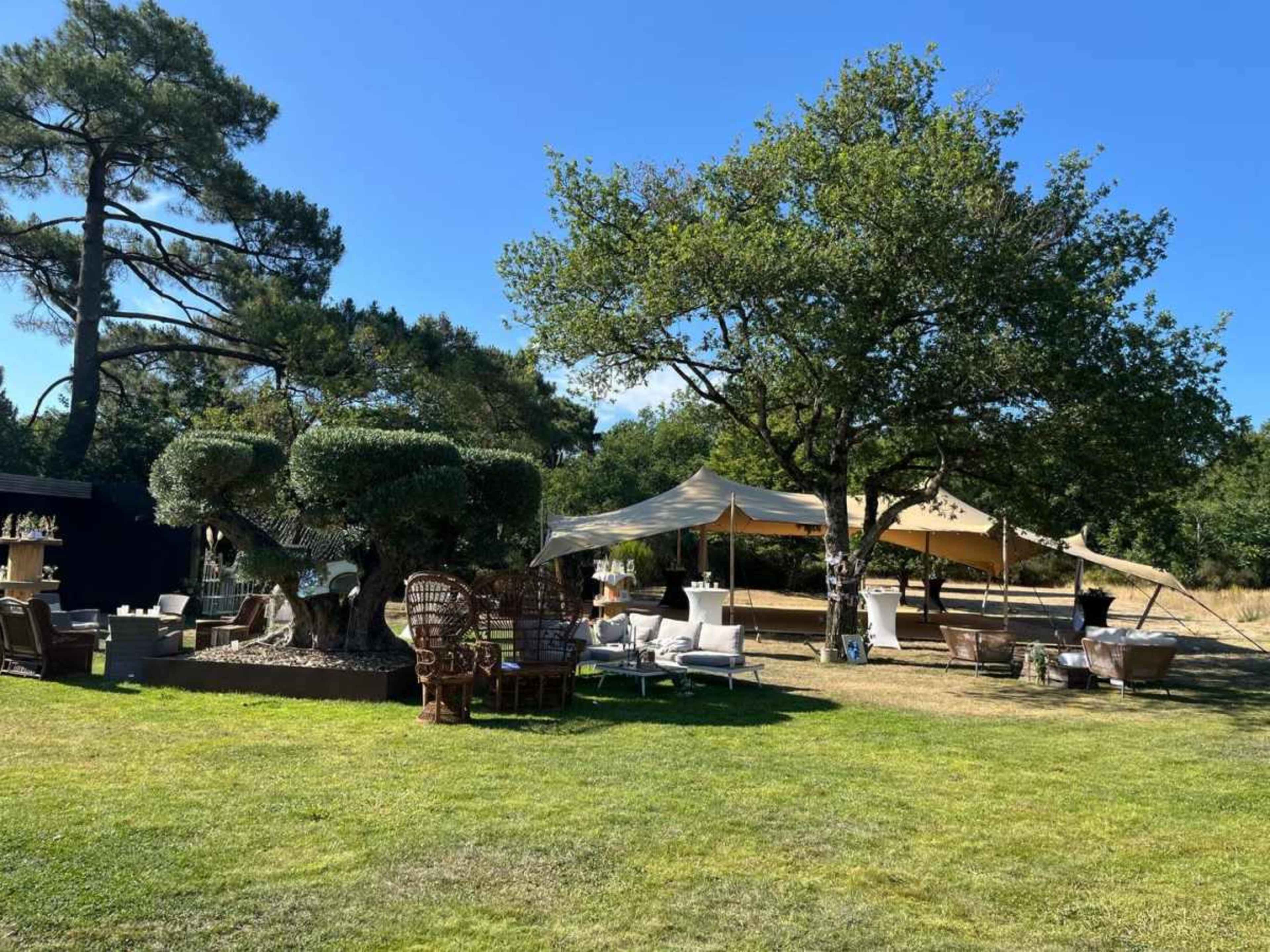 The scene shows a landscaped outdoor area with several seating arrangements, a large tent, trees, and manicured grass.