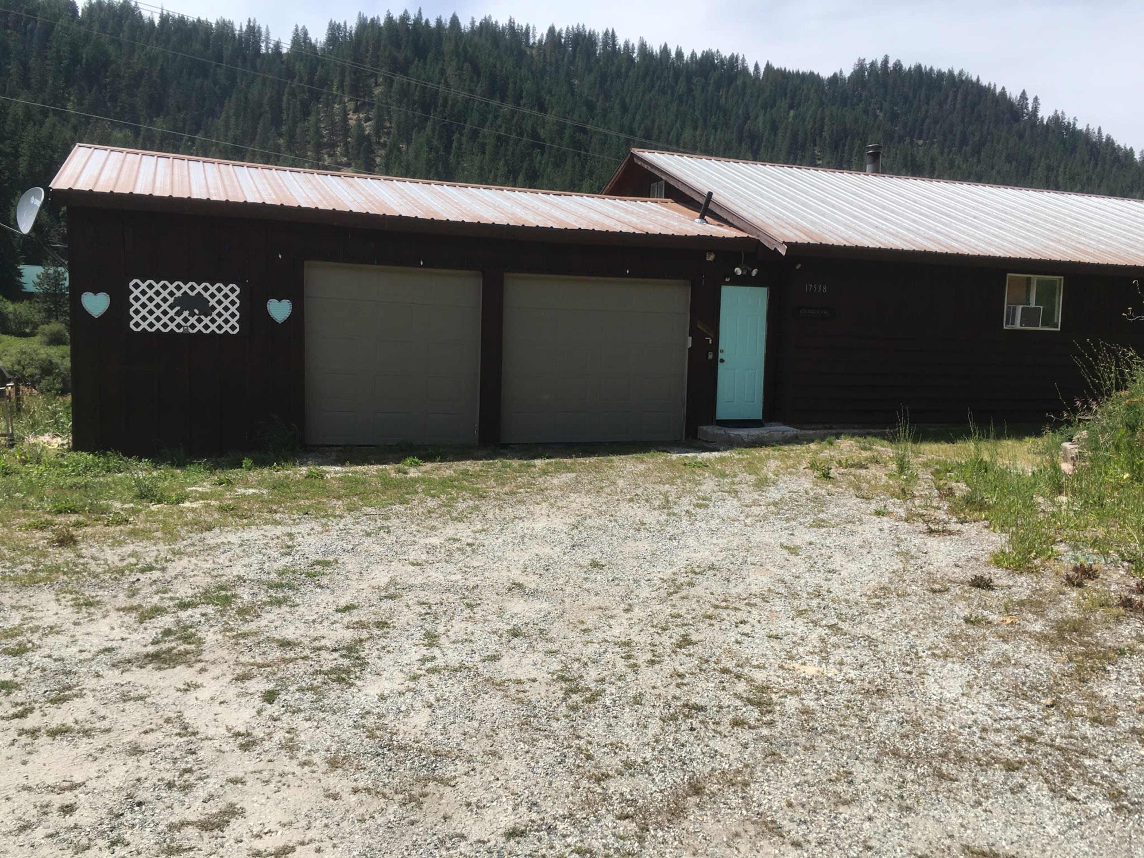 A dark wooden house with a metal roof and two garage doors, set on a gravel driveway in a mountainous area.