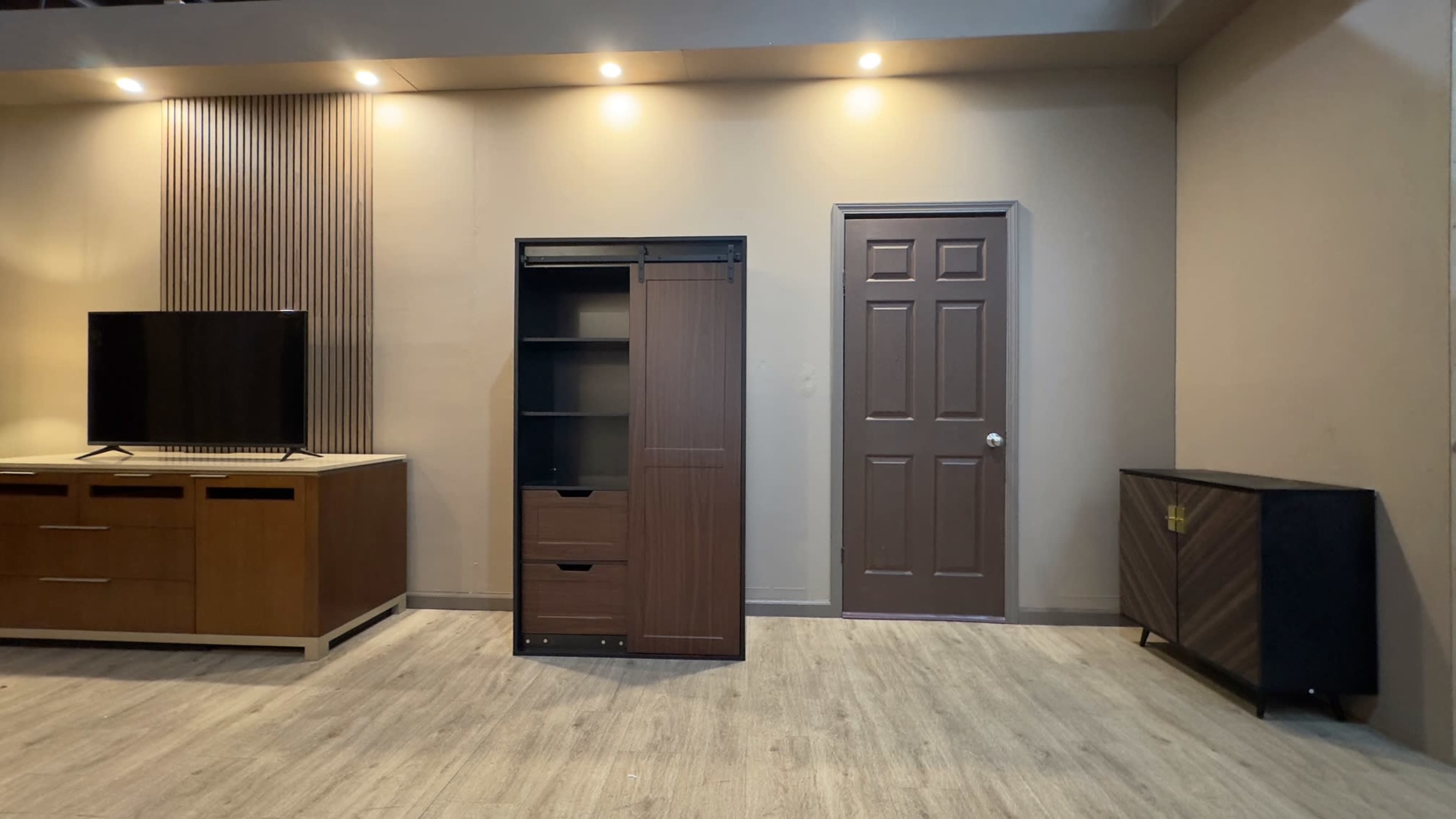 A modern interior space with a television on a wooden cabinet, a tall storage unit with drawers, a closed door, and a side cabinet against the wall.