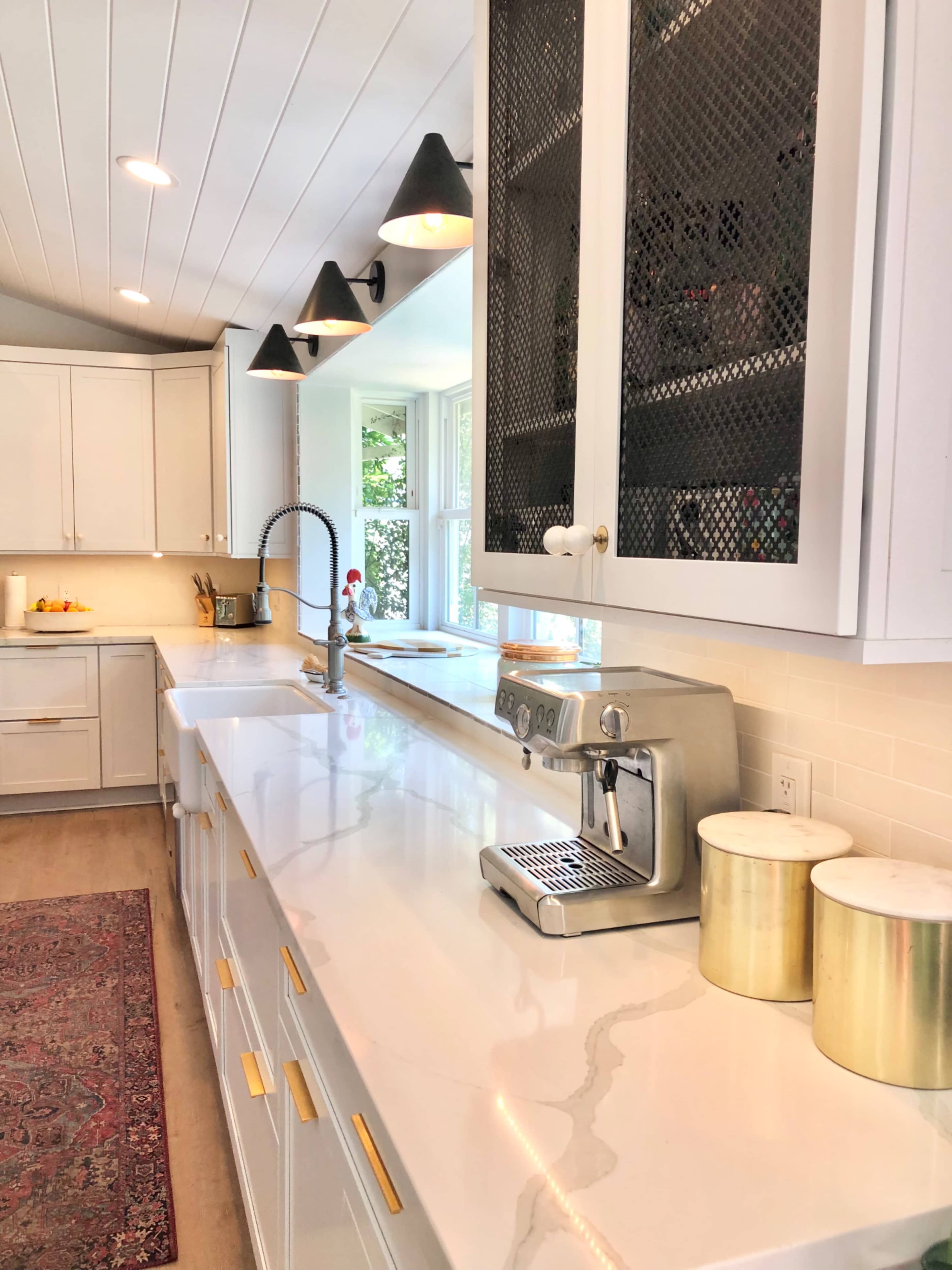 A modern kitchen features a central countertop with an espresso machine, white cabinetry, and a rug on the wooden floor.