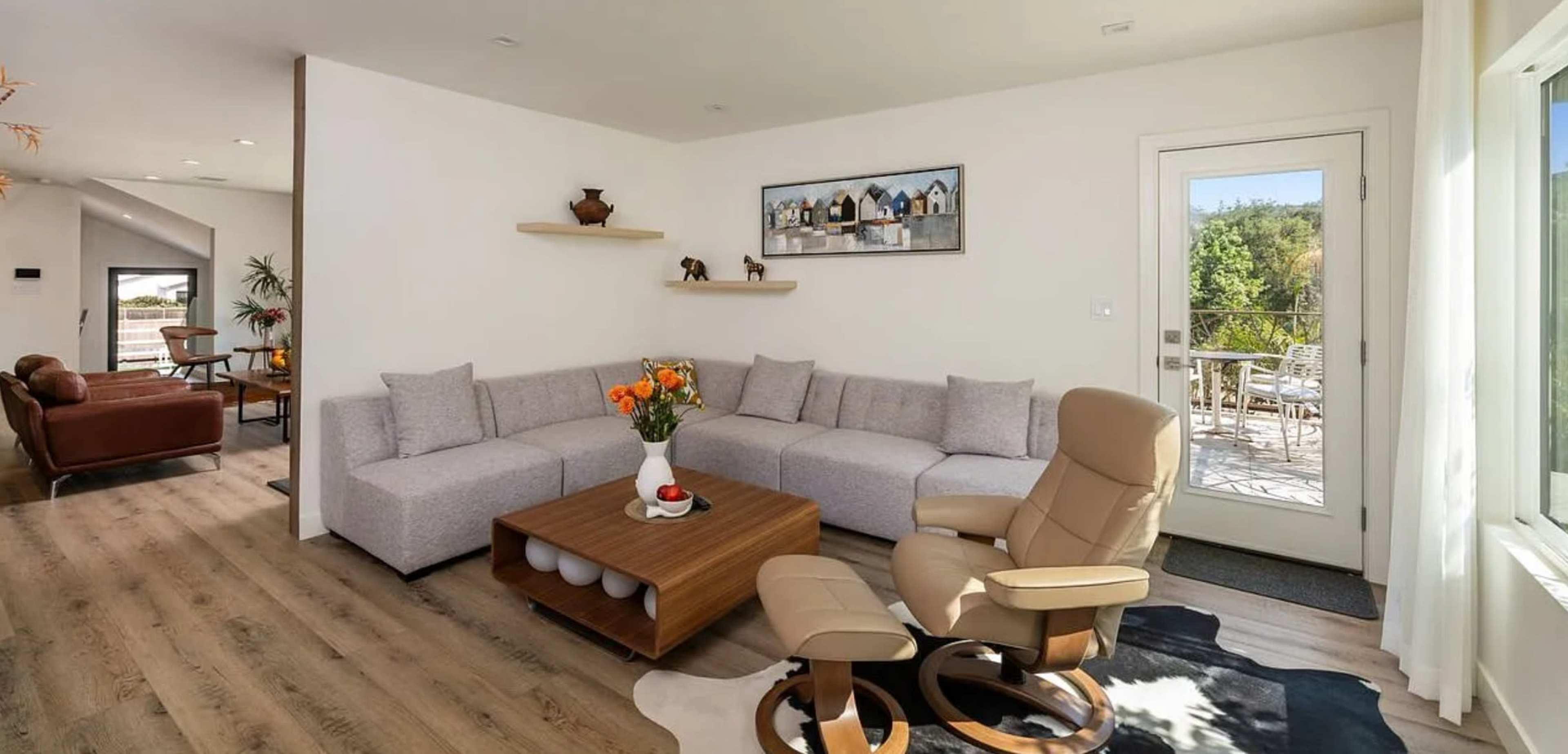 The image shows a modern living room with a sectional sofa, a wooden coffee table, and a doorway leading to a patio area.