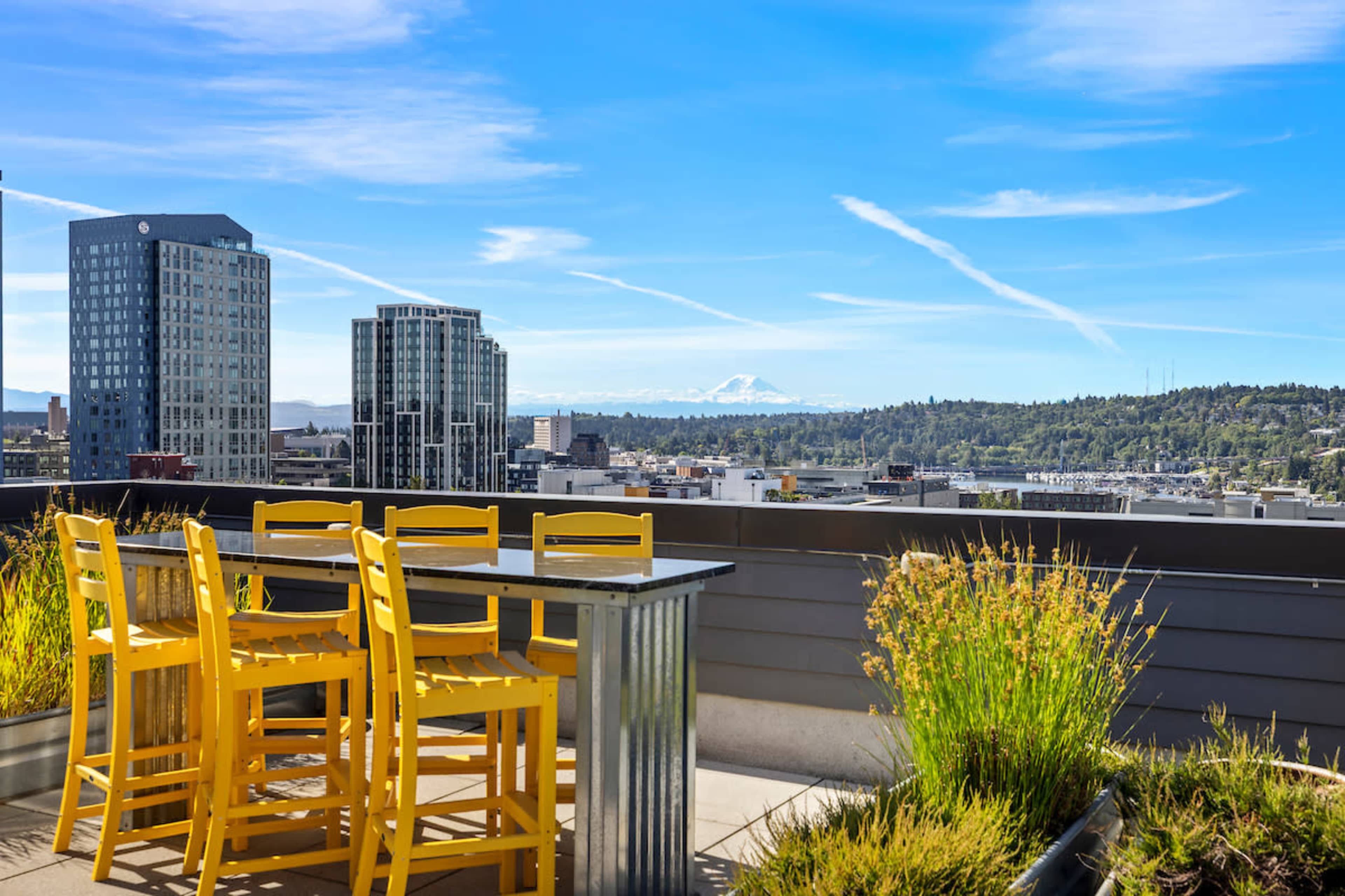 A rooftop terrace features a table and bright yellow chairs overlooking a city skyline with distant mountains under a clear blue sky.