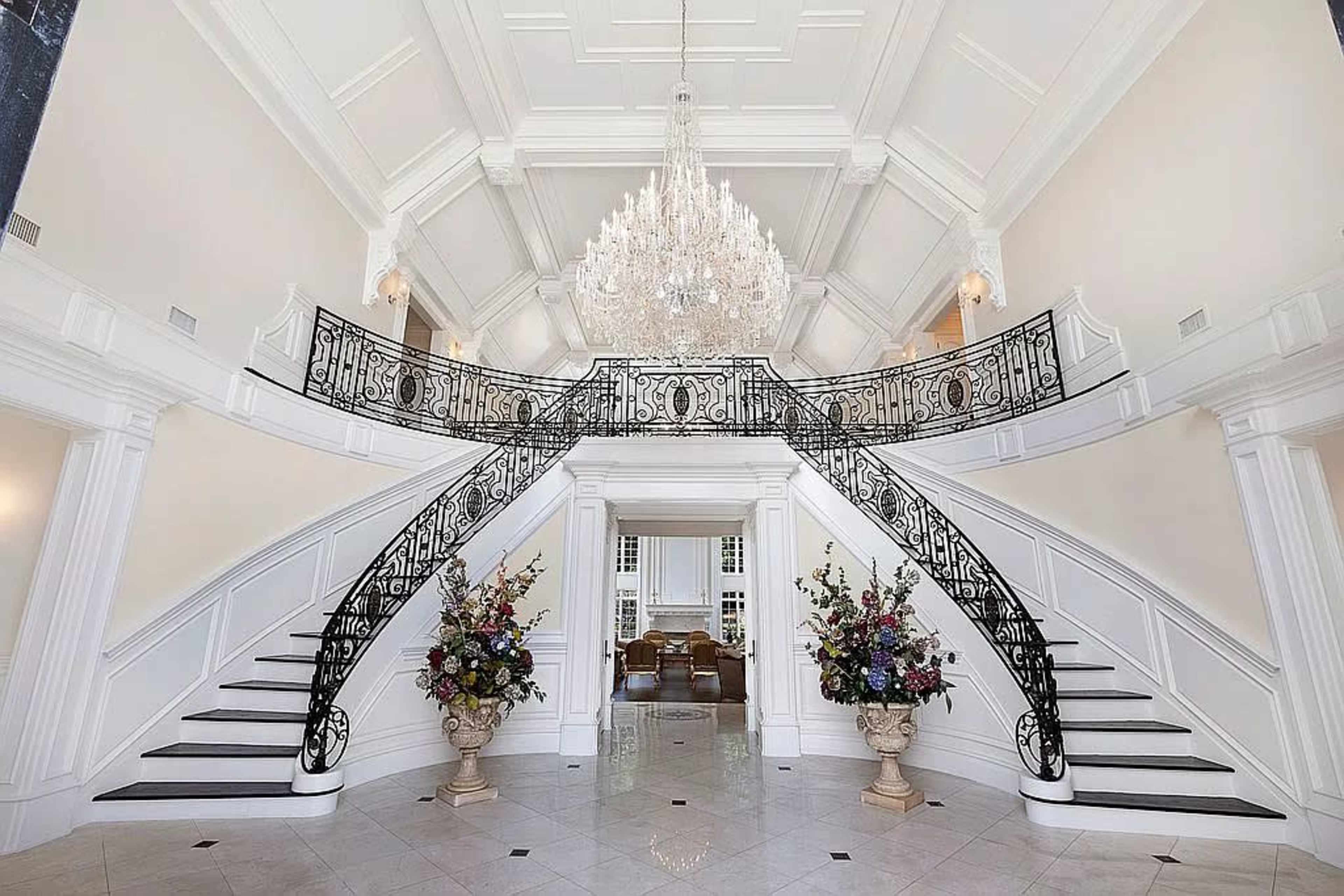 A grand foyer with a double staircase, ornate iron railings, a large chandelier, and decorative floral arrangements in marble vases.