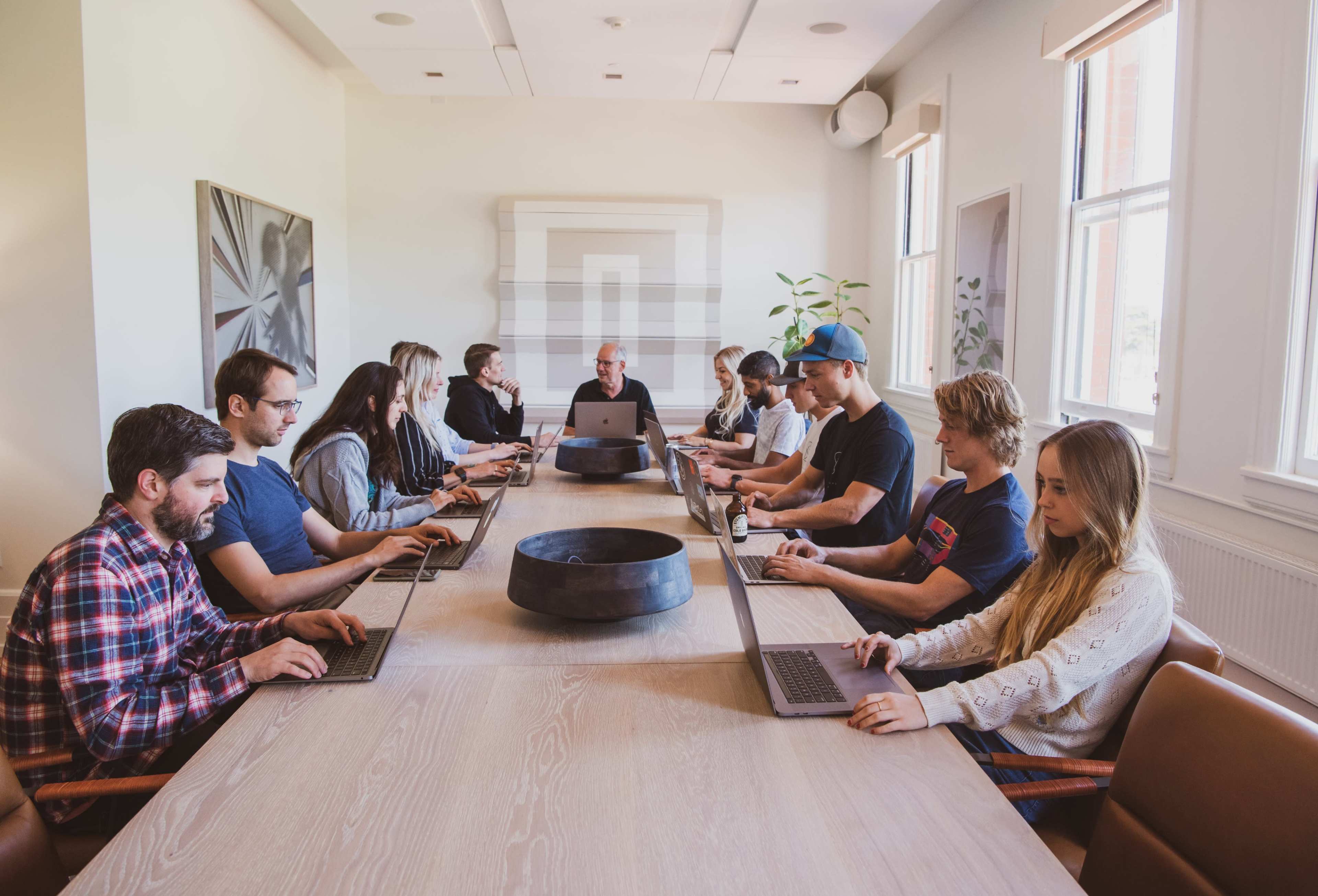 A group of twelve people are seated at a long table in a bright conference room, each working on their laptops.