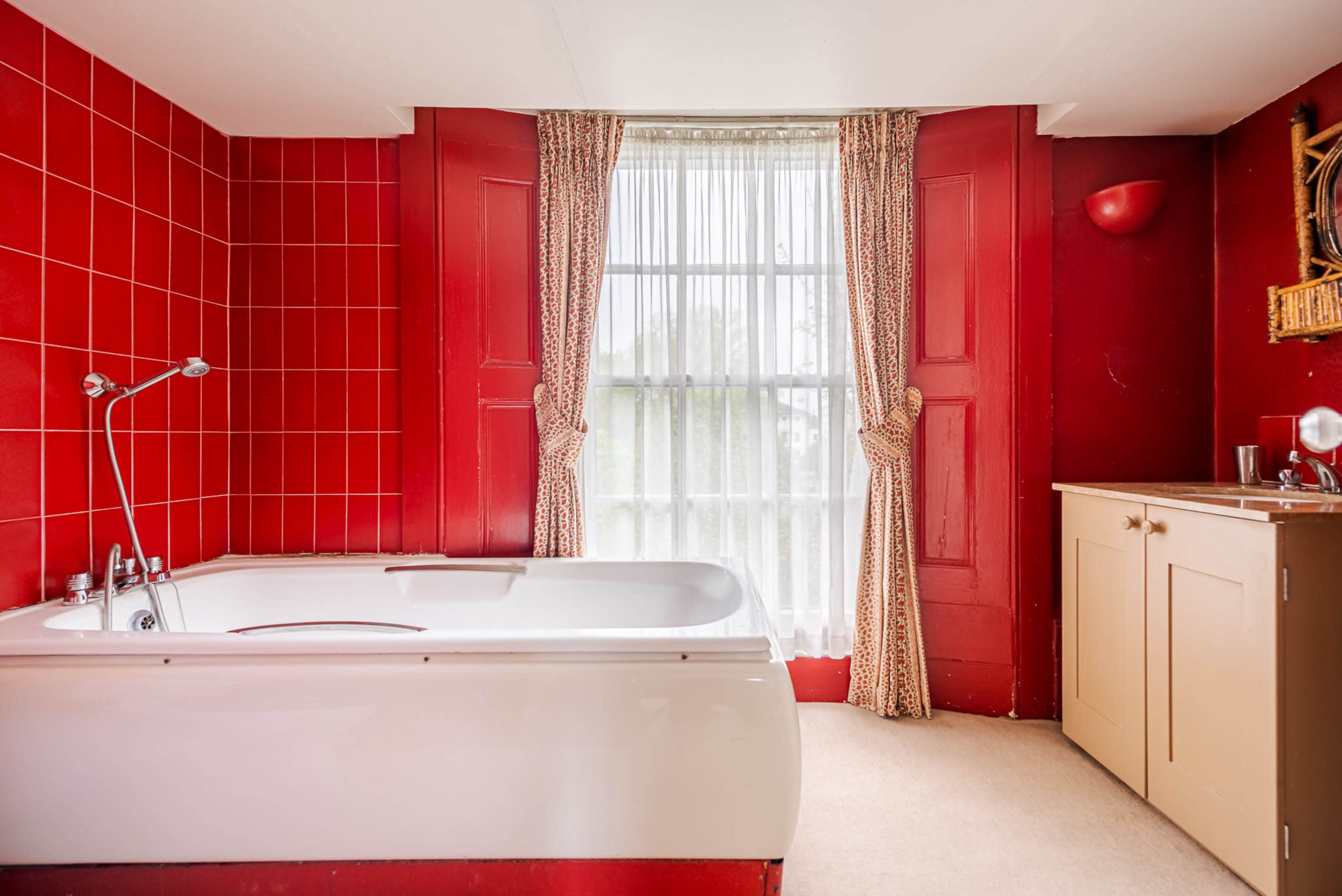 A bathroom features a freestanding white bathtub against a backdrop of red tiled walls and a large window with patterned curtains.