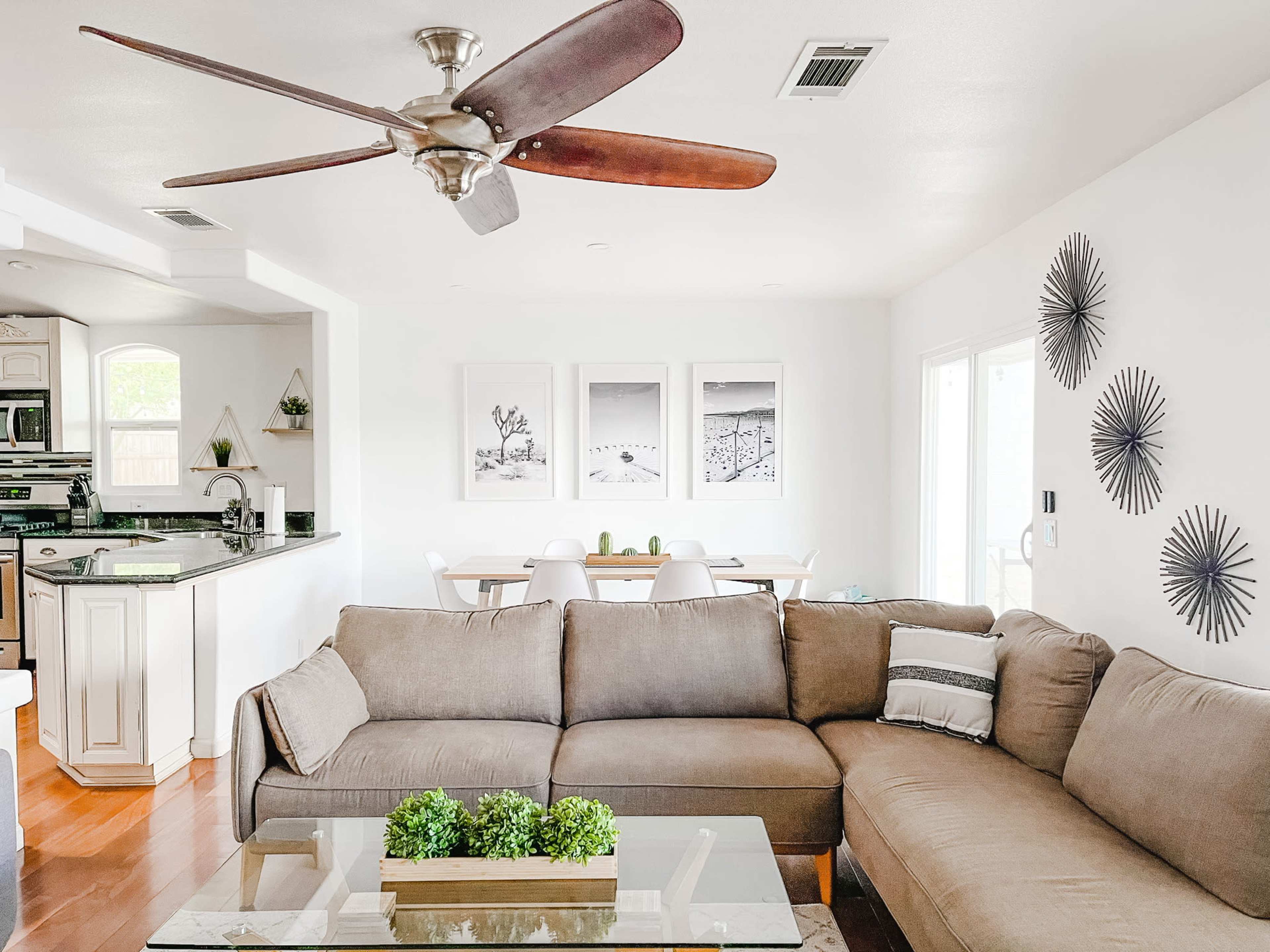 A bright living room features a sectional sofa, a glass coffee table with greenery, and a ceiling fan overhead, with a dining area visible in the background.