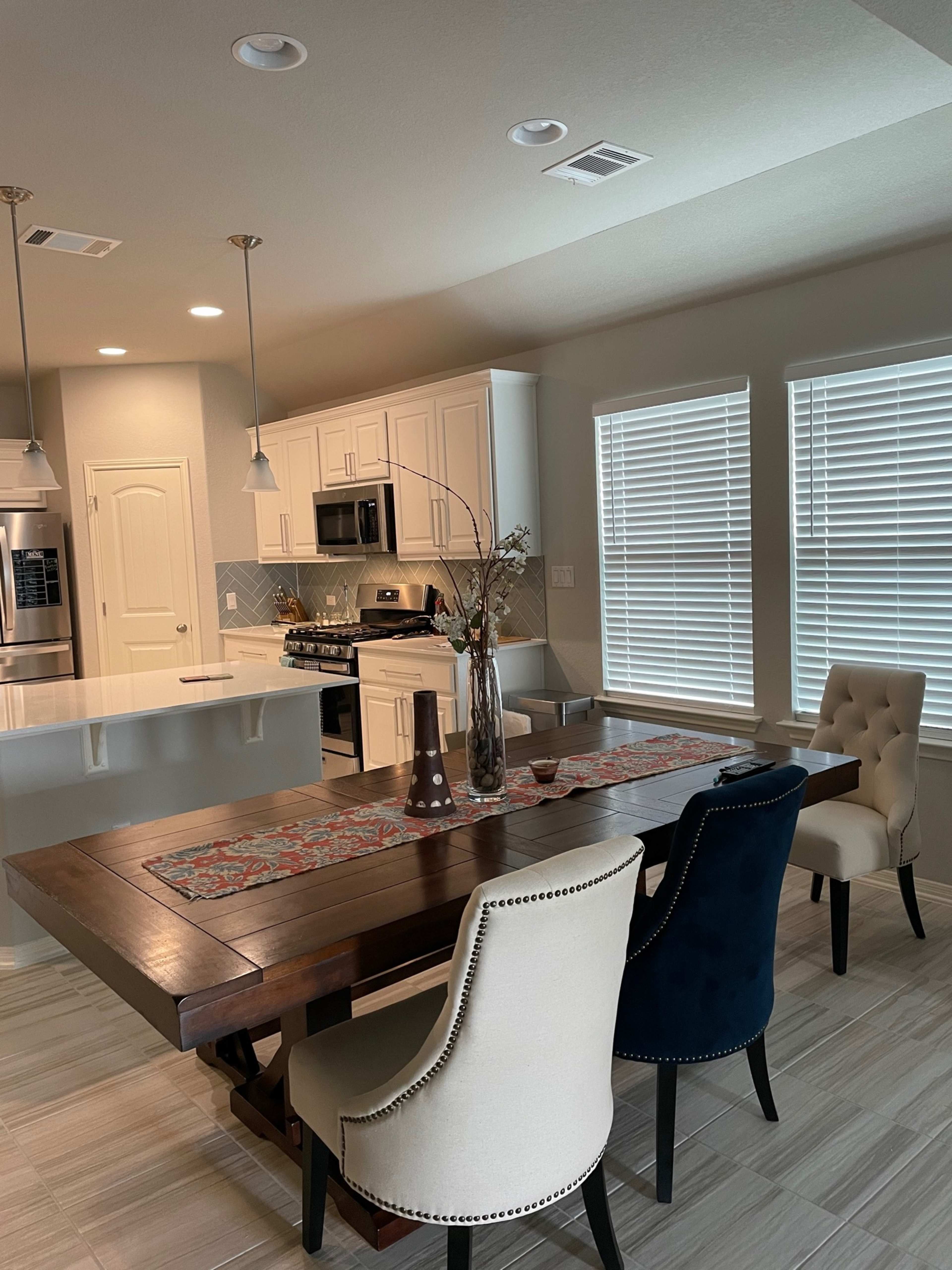 The image shows a modern kitchen and dining area featuring a wooden dining table with two upholstered chairs, a gray-colored kitchen with white cabinets, and stainless steel appliances.