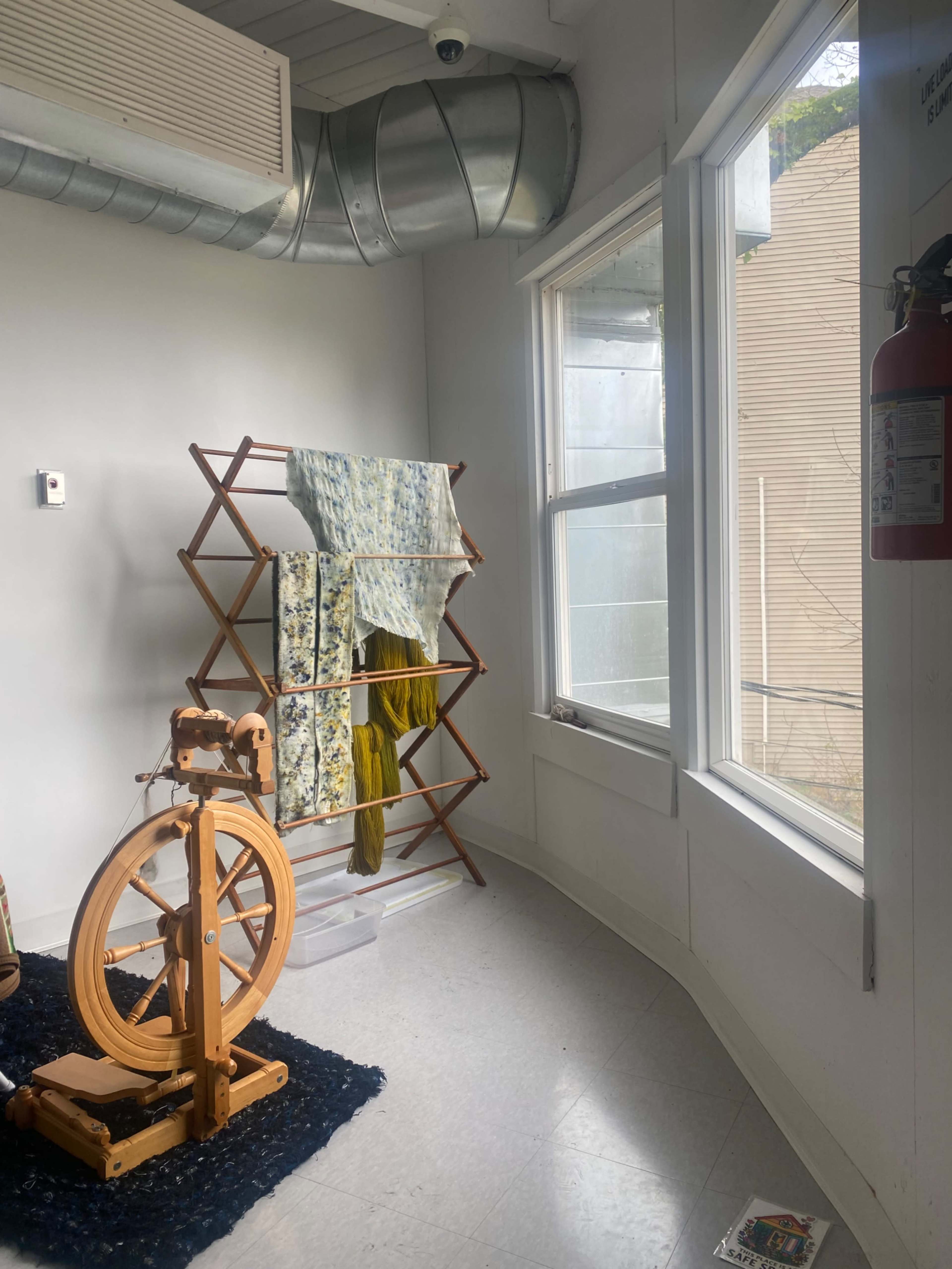 The image shows a light-colored room featuring a wooden drying rack with clothes, a spinning wheel, and large windows allowing natural light to enter.