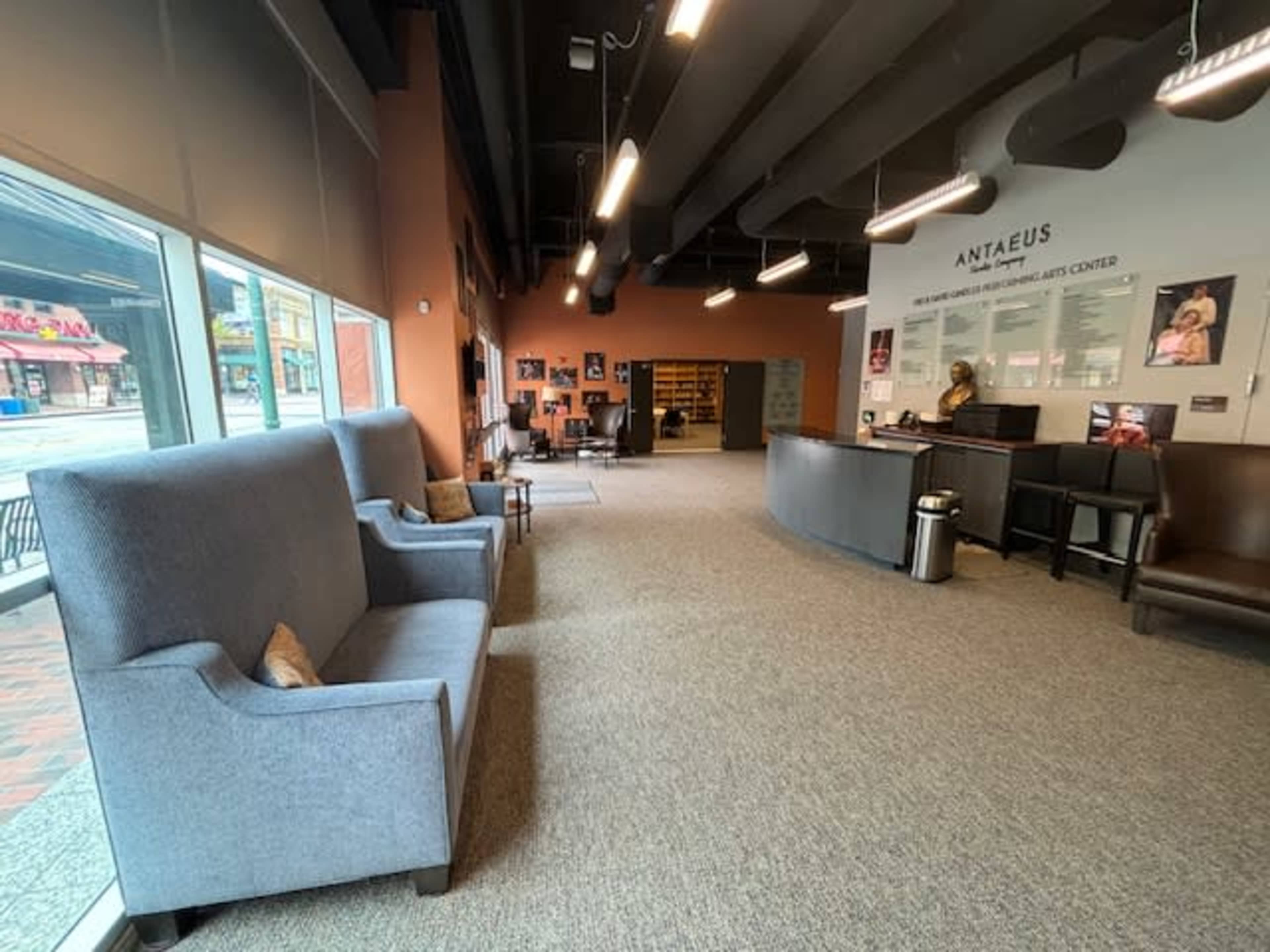 The image shows a spacious lobby area of the Antaeus Theatre, featuring two gray armchairs, a reception desk, and walls adorned with posters and information panels.