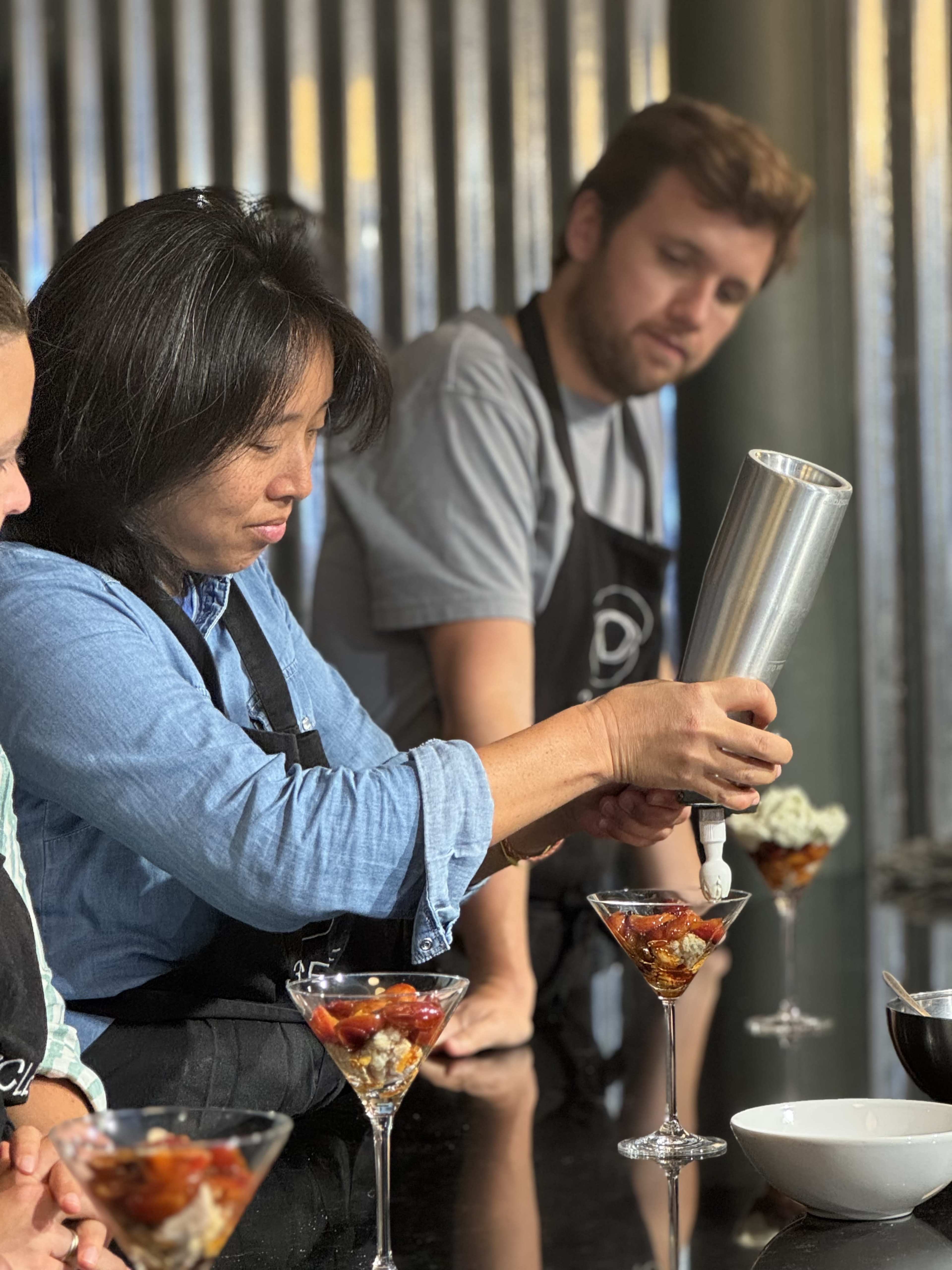 A woman in an apron carefully pipes a garnish onto dessert-filled glasses while a man in a gray shirt observes in the background.