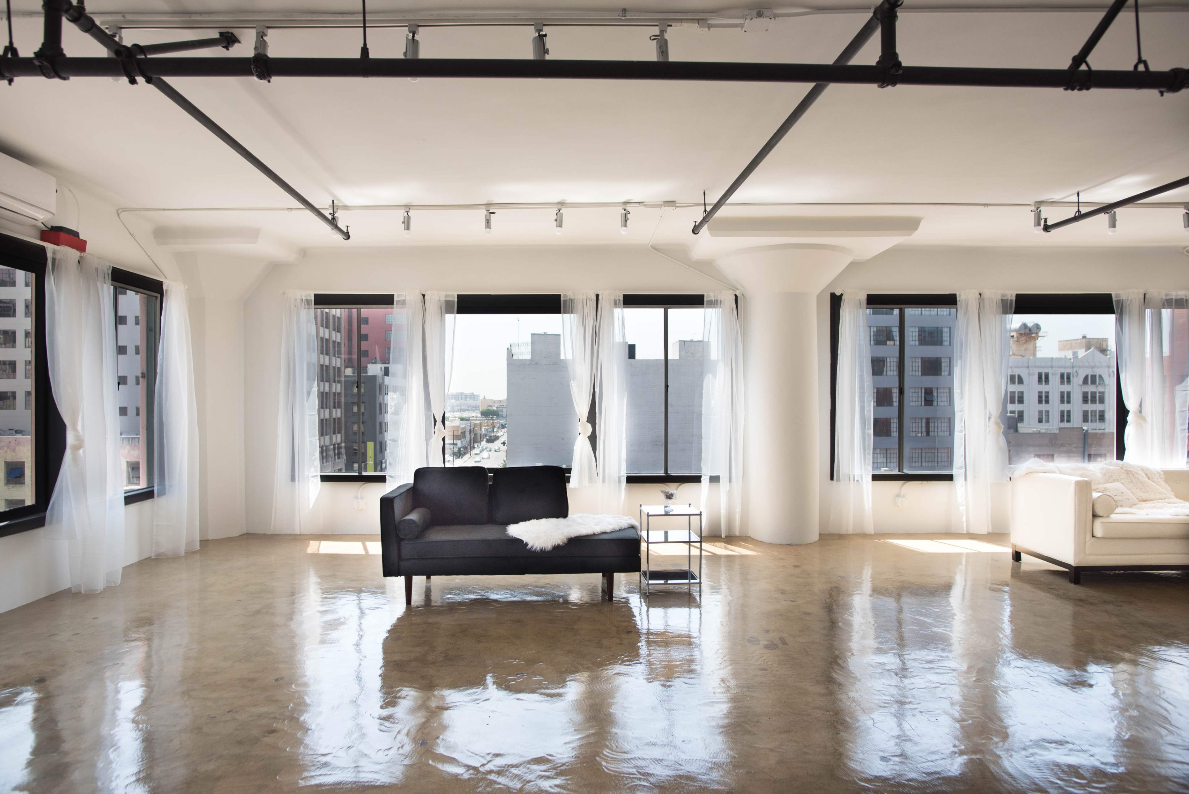 A spacious, modern room with large windows, a black sofa, a light-colored armchair, and polished concrete floors.
