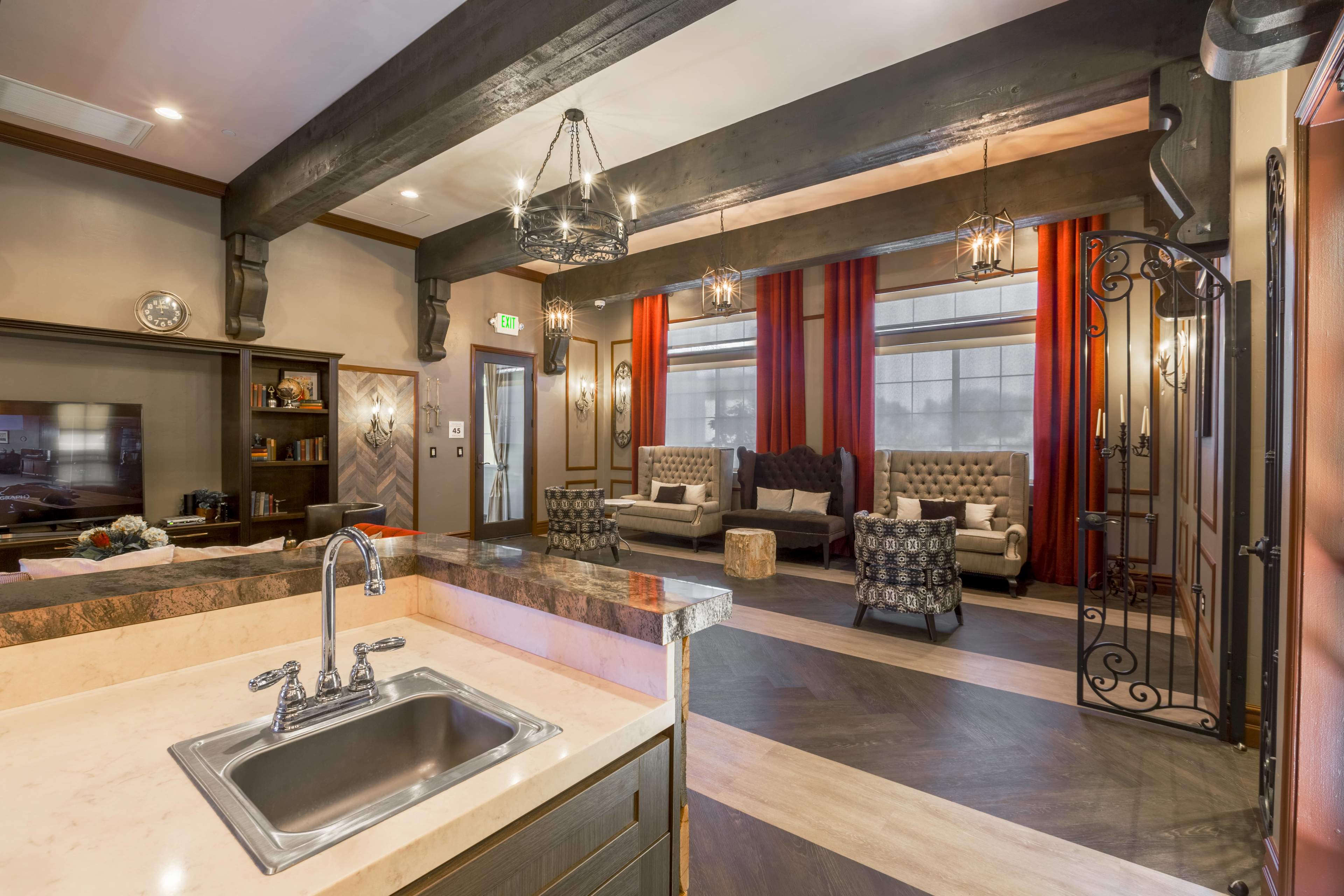 The image shows a modern living area with a kitchen counter featuring a sink, dark wooden beams above, red curtains, and stylish seating arrangements.
