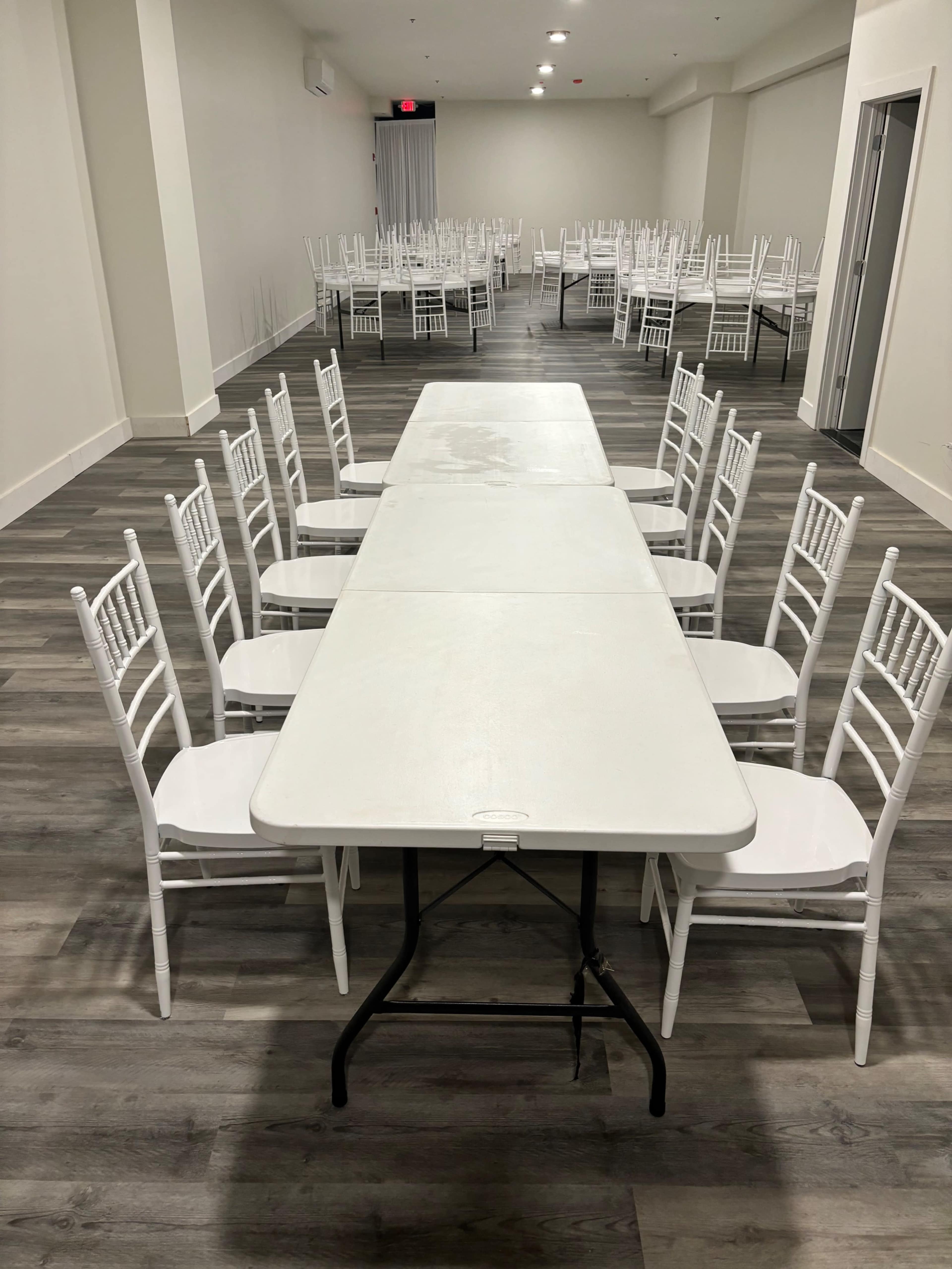 A long white table is set up with white chairs on either side in a spacious, empty room.