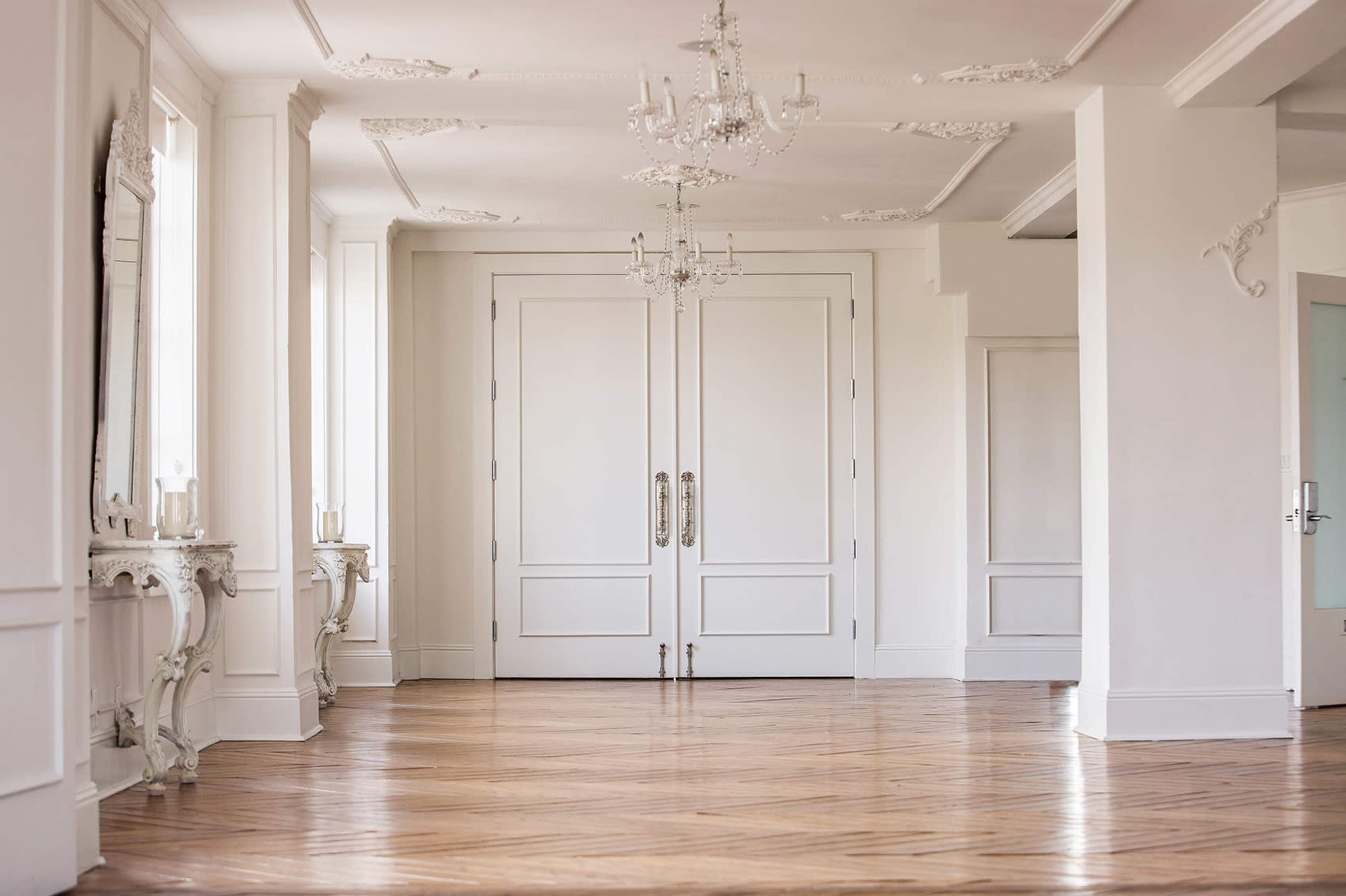 The image shows a spacious interior room with hardwood floors, large white double doors, and ornate chandeliers.