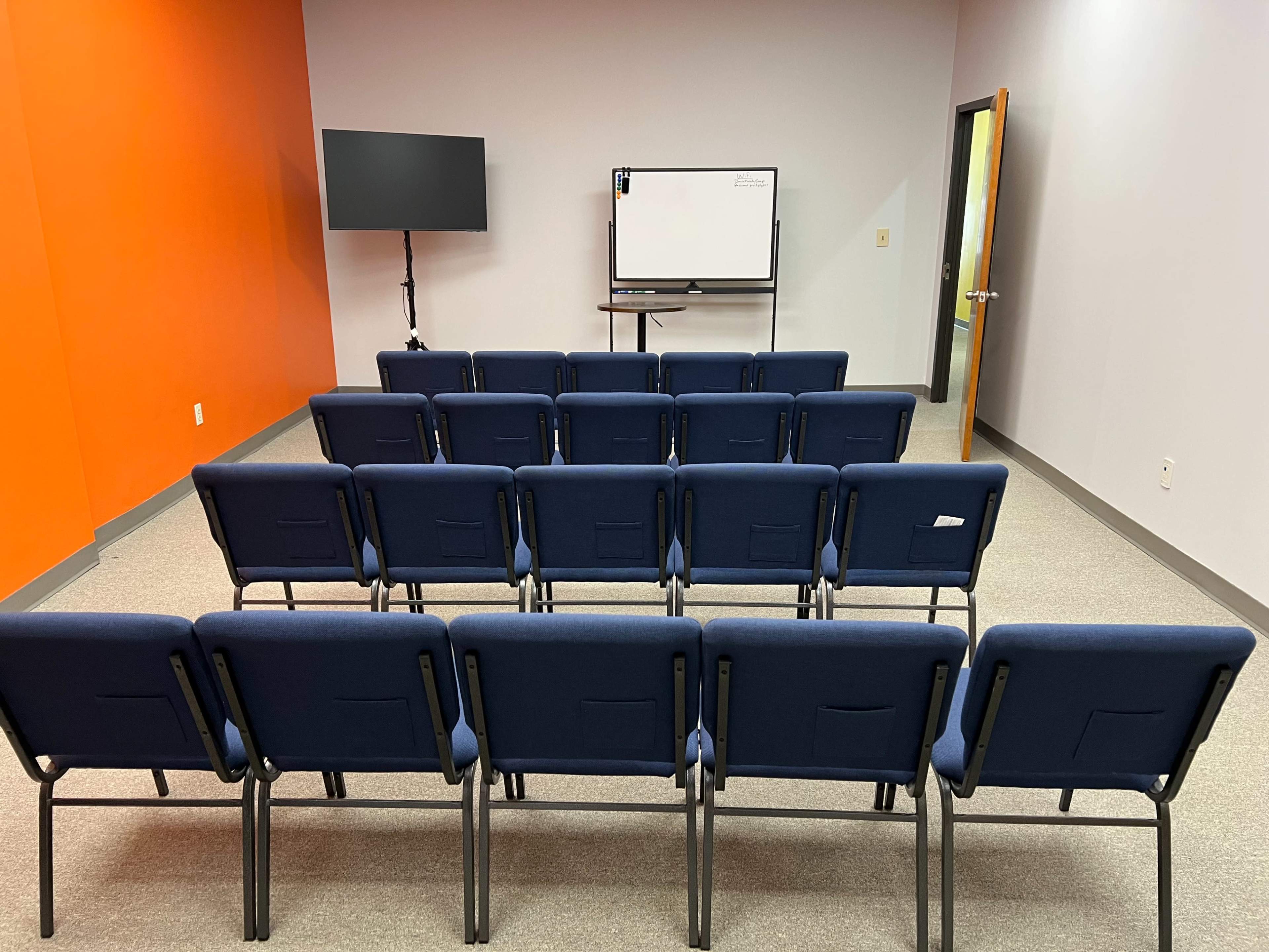 The image shows a classroom or meeting room with rows of blue chairs facing a whiteboard and a monitor on a stand, against an orange wall.