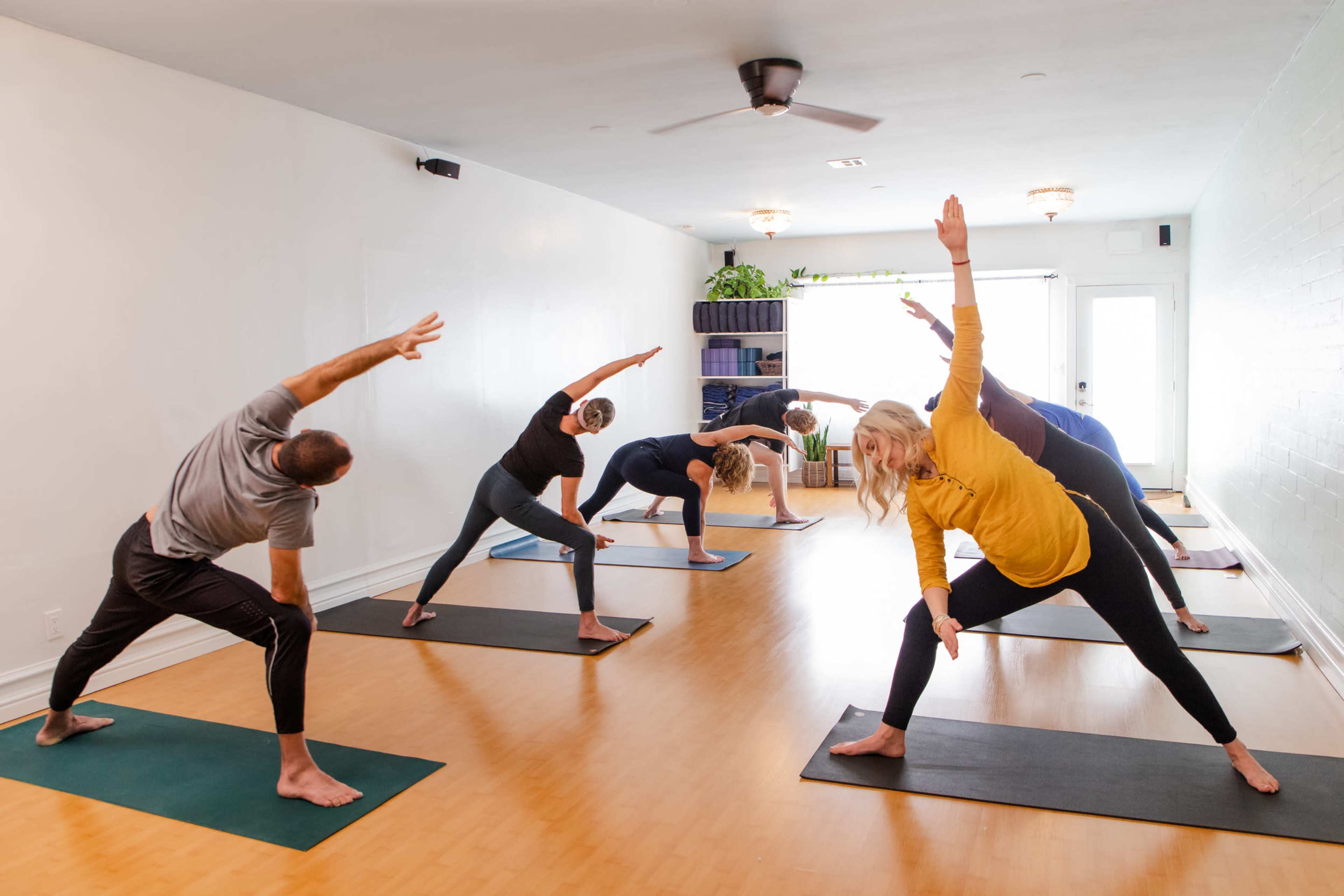 A group of individuals is practicing yoga in a well-lit studio, each person engaged in a different pose on green mats.