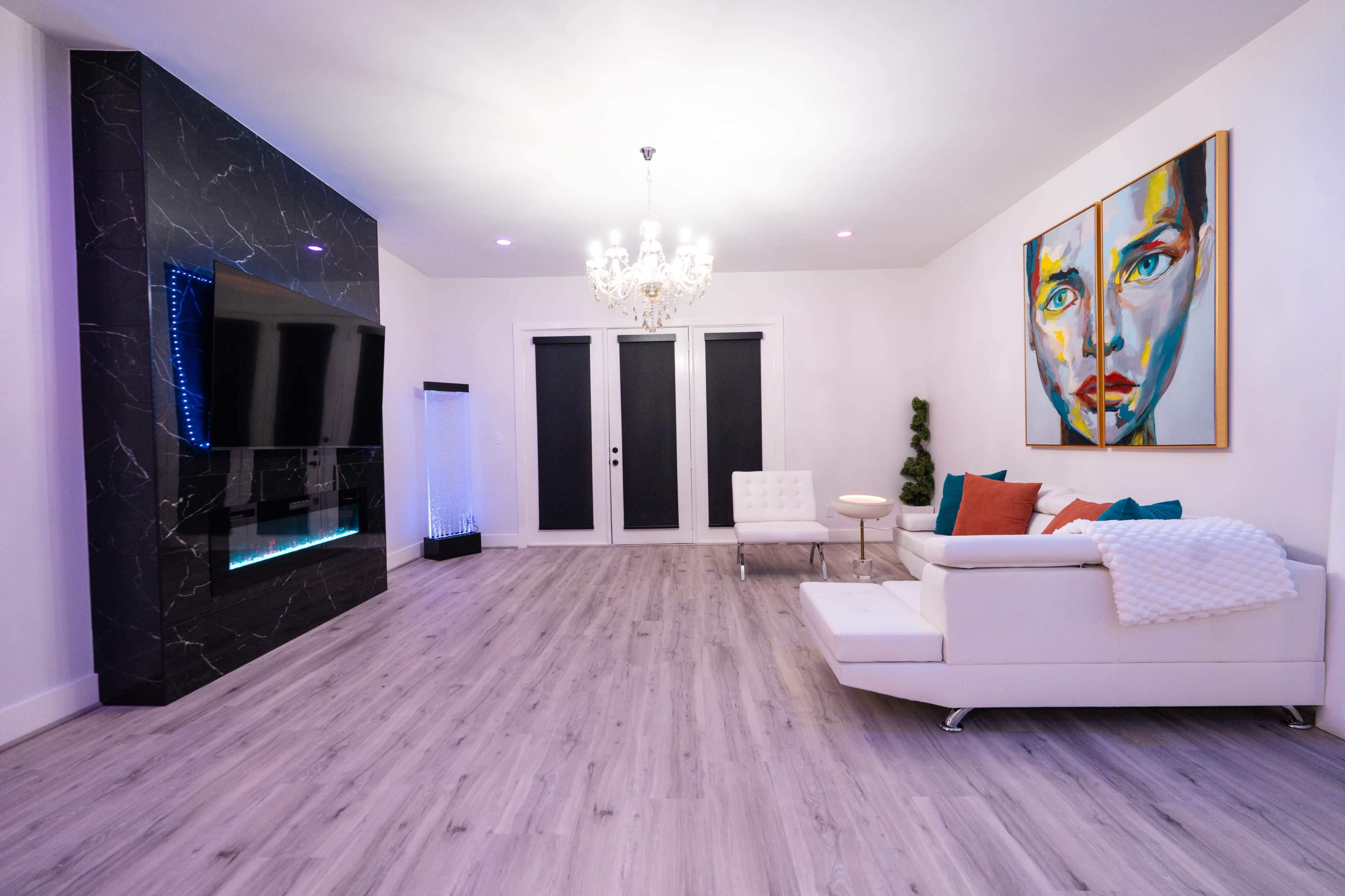 A modern living room features a white sofa, a black and white marble accent wall with a TV, colorful artwork, and a chandelier above.