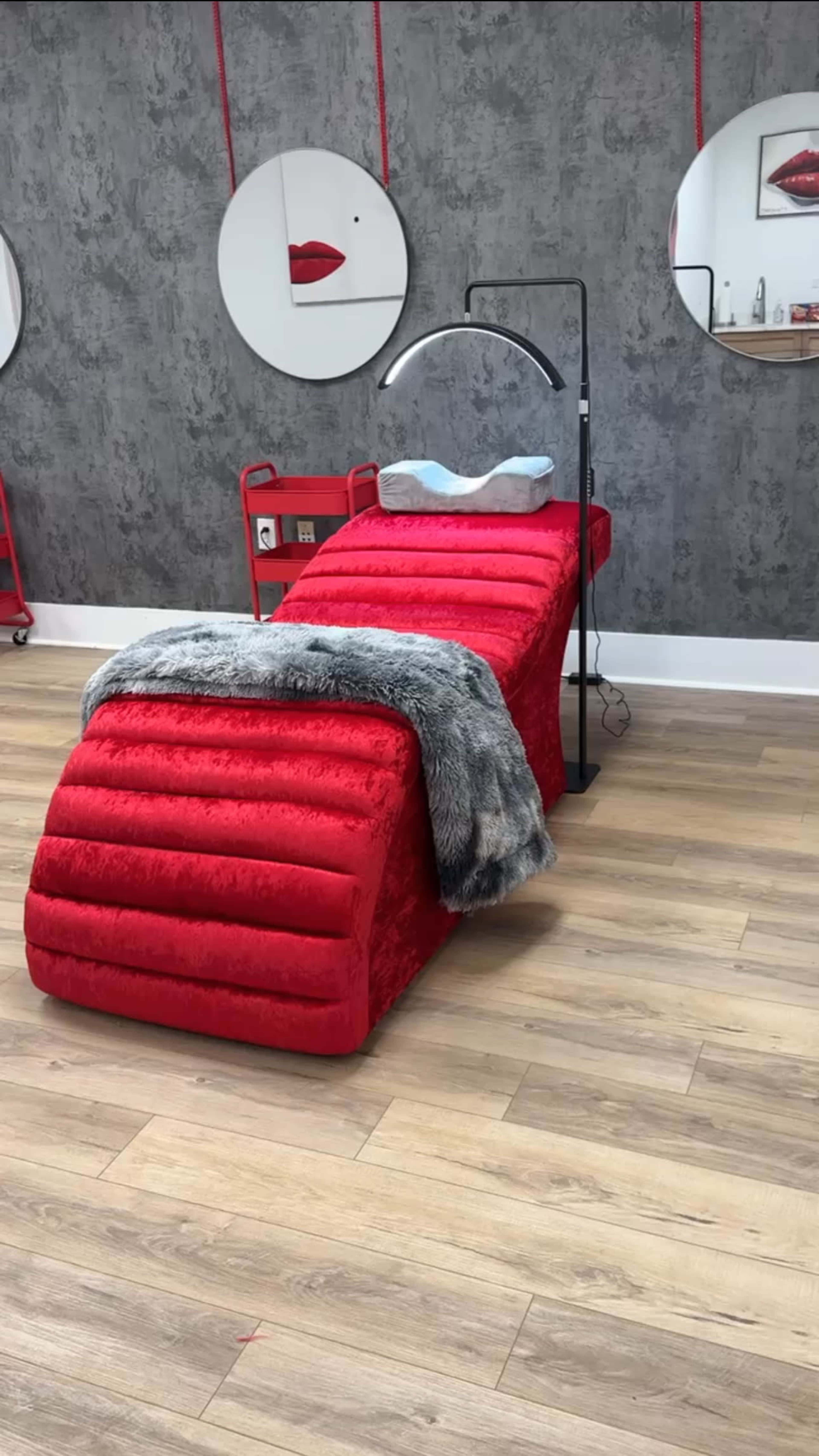 The image shows a red treatment bed with a gray throw blanket and a lamp next to it, set against a textured wall and wooden floor.