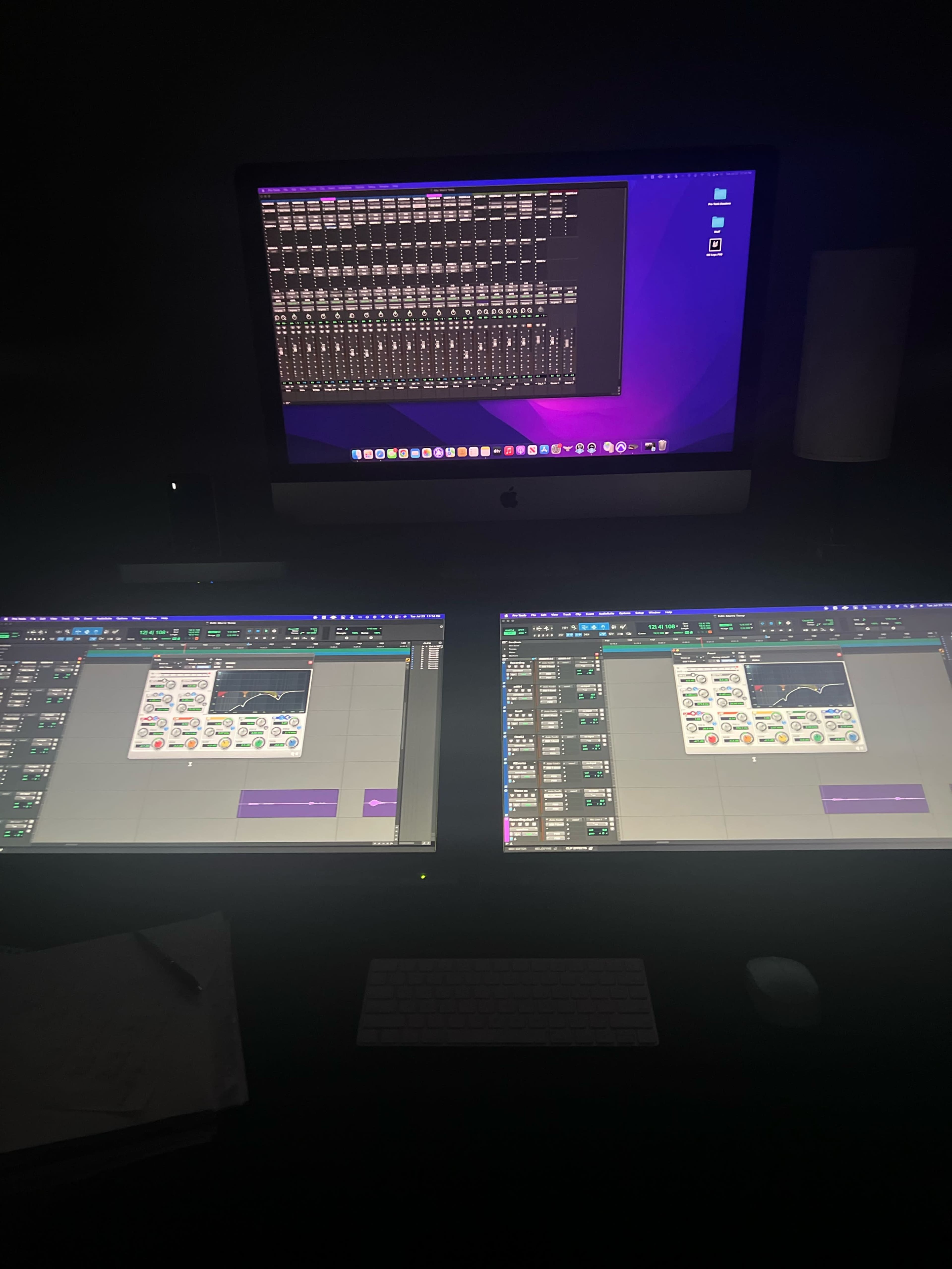 The image shows a dimly lit workspace with three computer screens displaying audio editing software and a mixing interface.