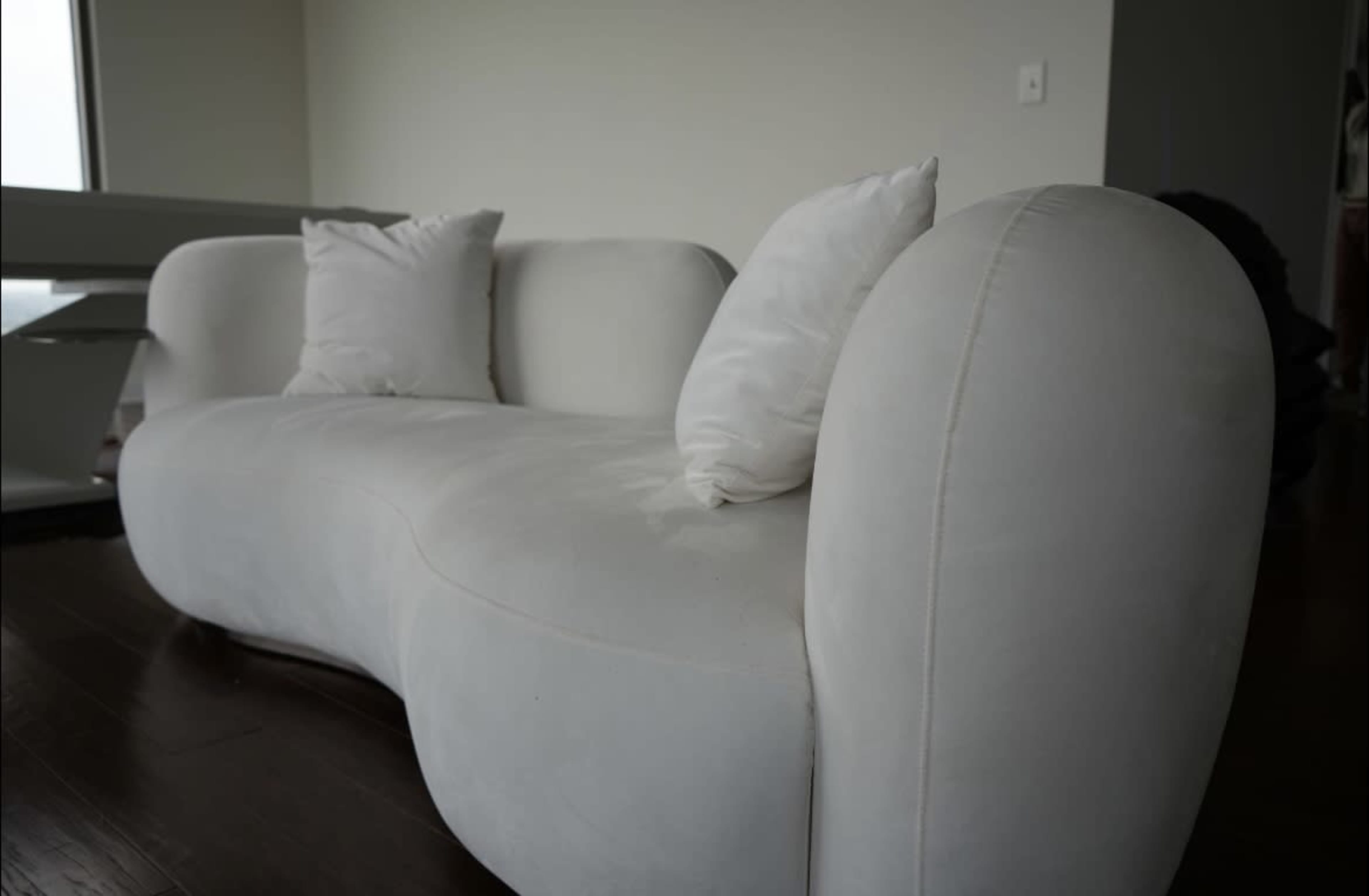 A white, curved sofa with two cushions sits on a dark wooden floor near a large window.