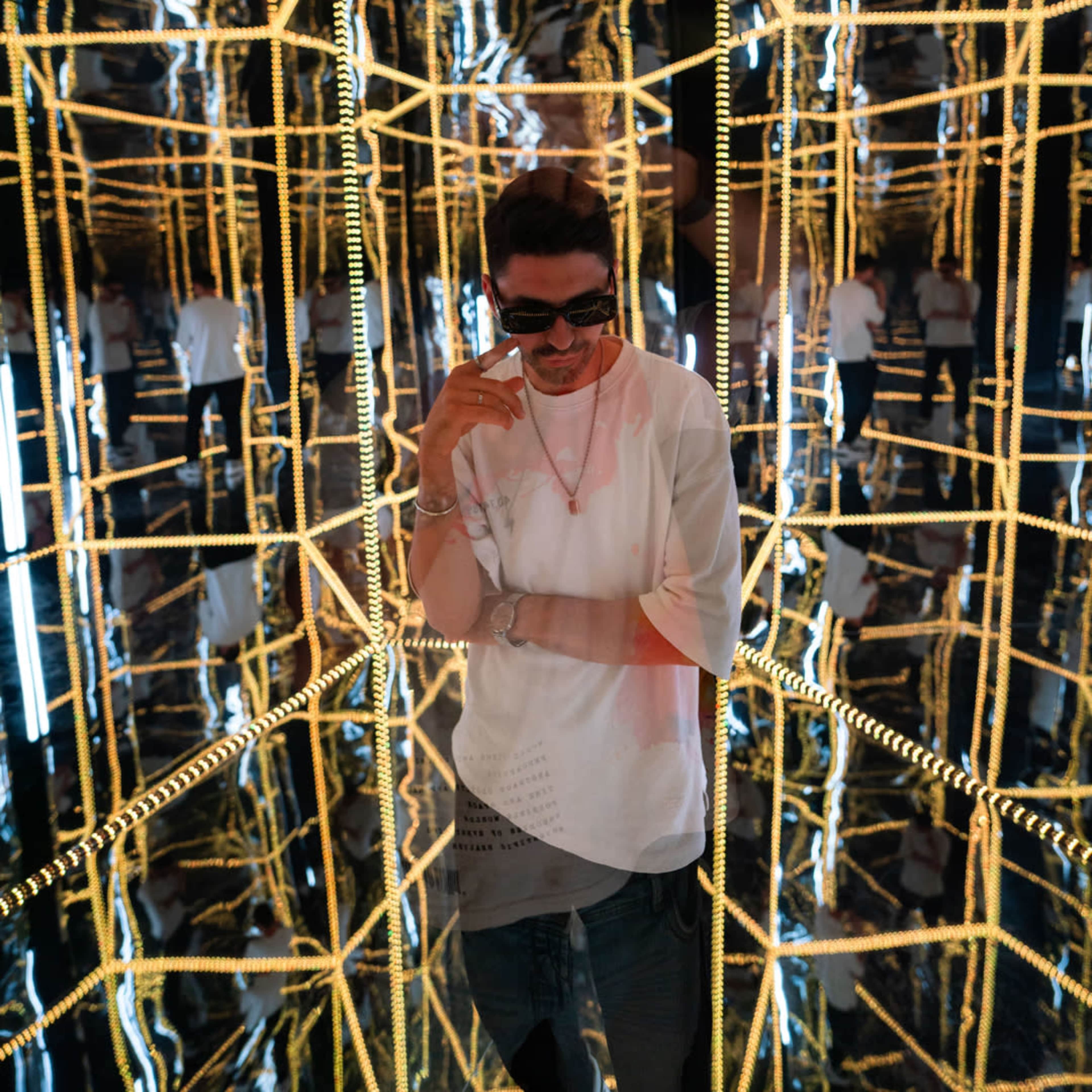 A person wearing sunglasses stands in a mirrored room illuminated by yellow lights, creating multiple reflections.