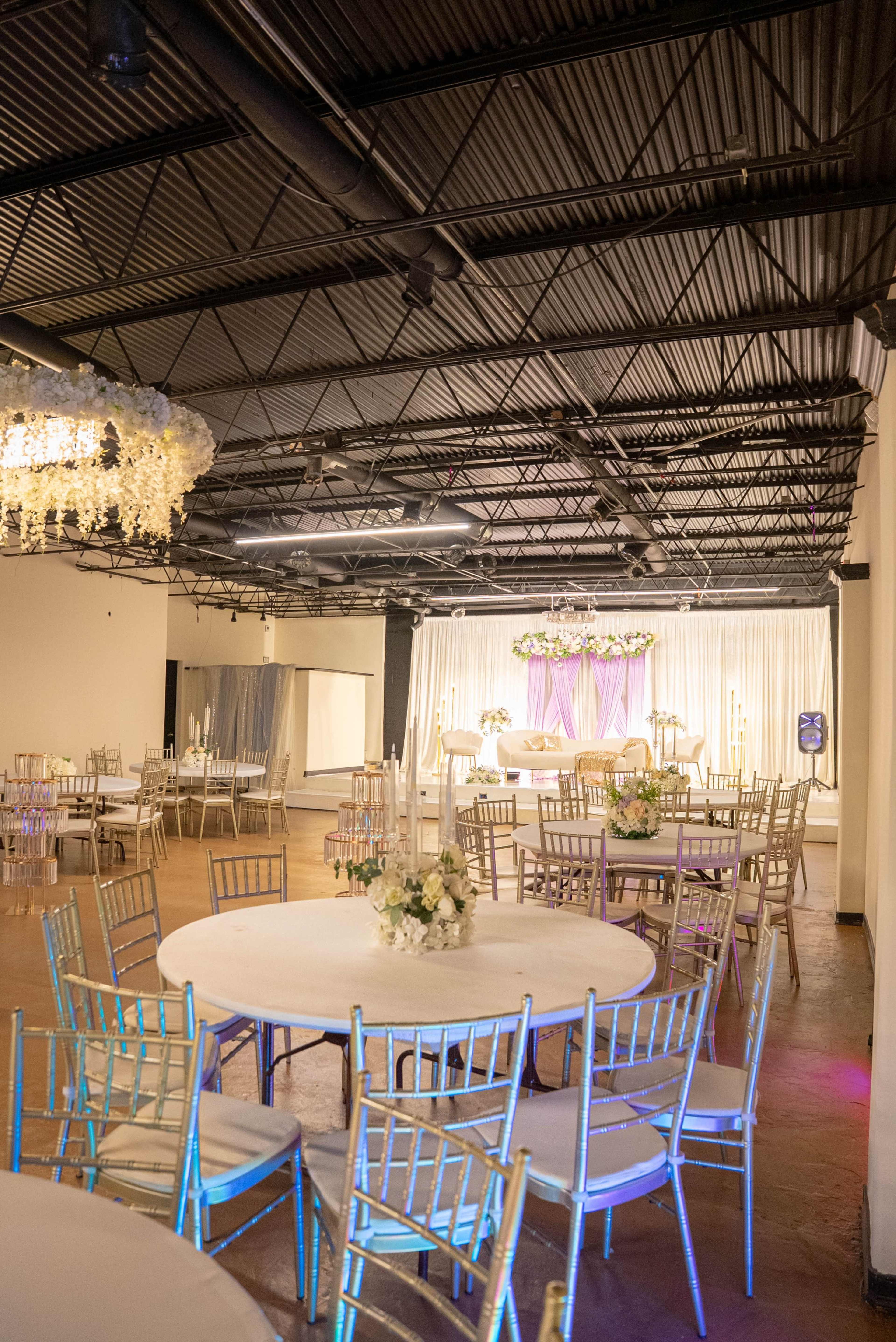 The image shows a spacious event venue with round tables set up, decorated with floral centerpieces, and a stage area adorned with drapes and floral arrangements.