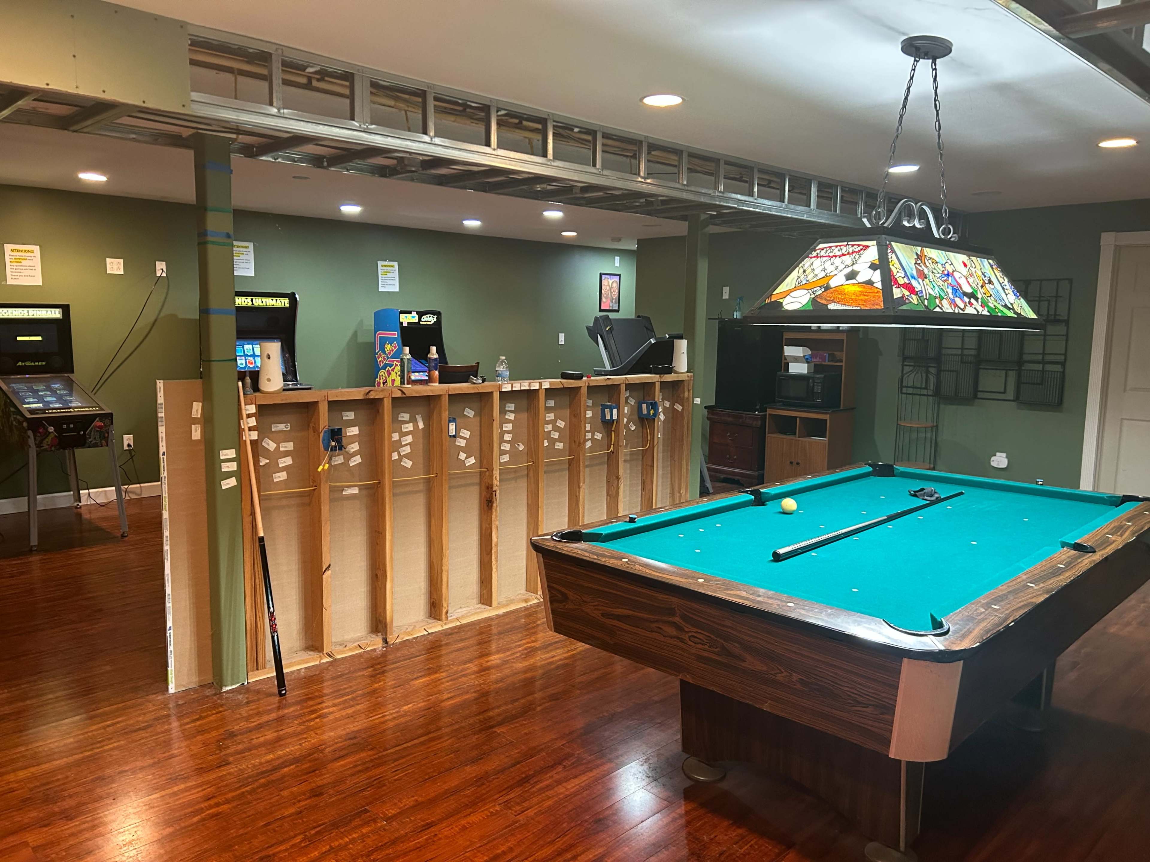 The image shows a basement game room featuring a pool table, a bar-like counter with various machines, and a green-painted wall.