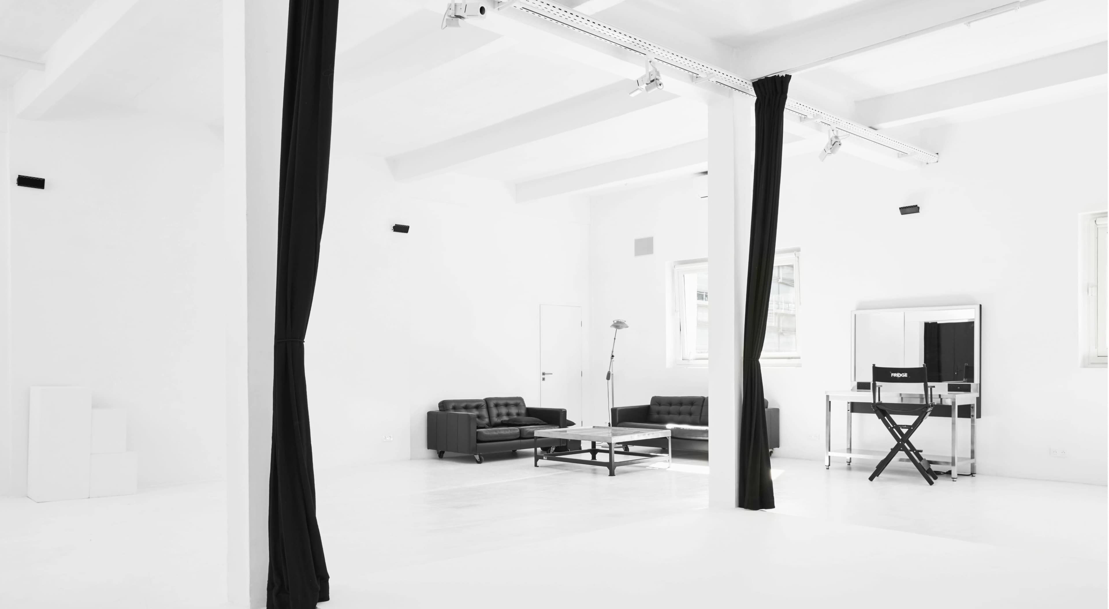 A minimalist, white room with black curtains, a black couch, a coffee table, and a mirrored dressing table.