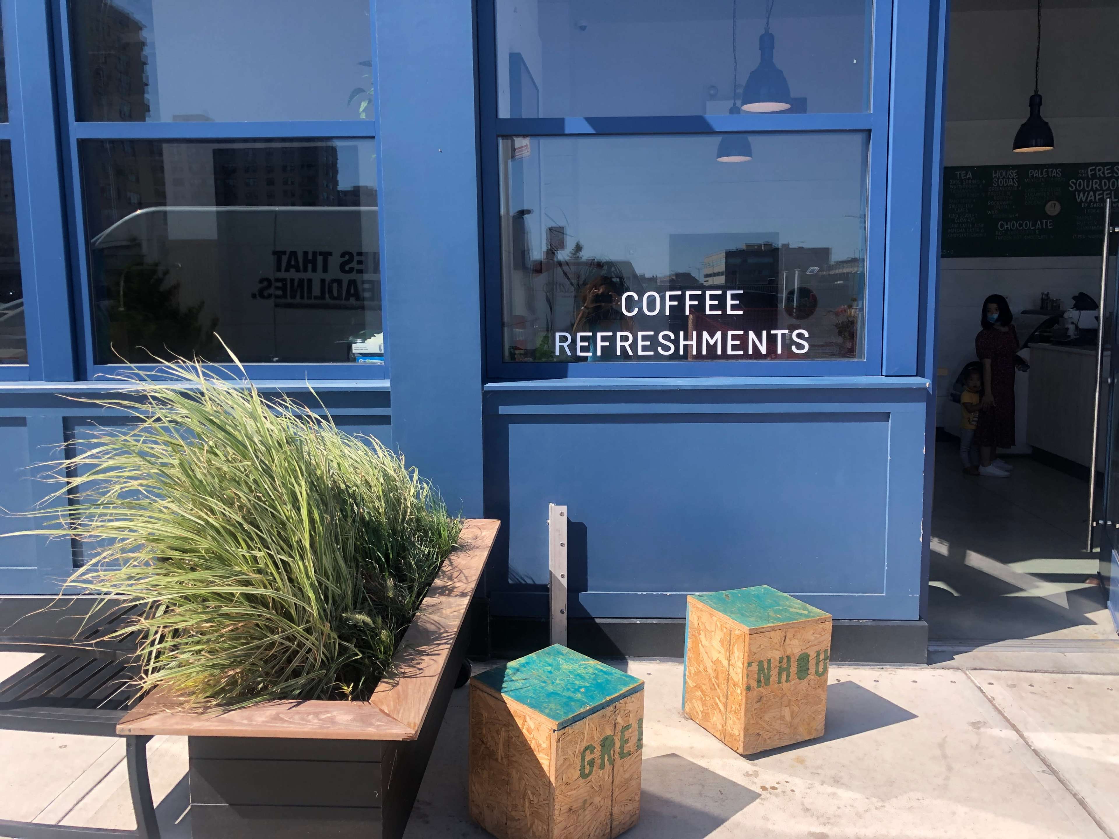 A blue building with large windows displays signs for "Coffee" and "Refreshments," while two wooden stools are placed outside near a planter of grass.
