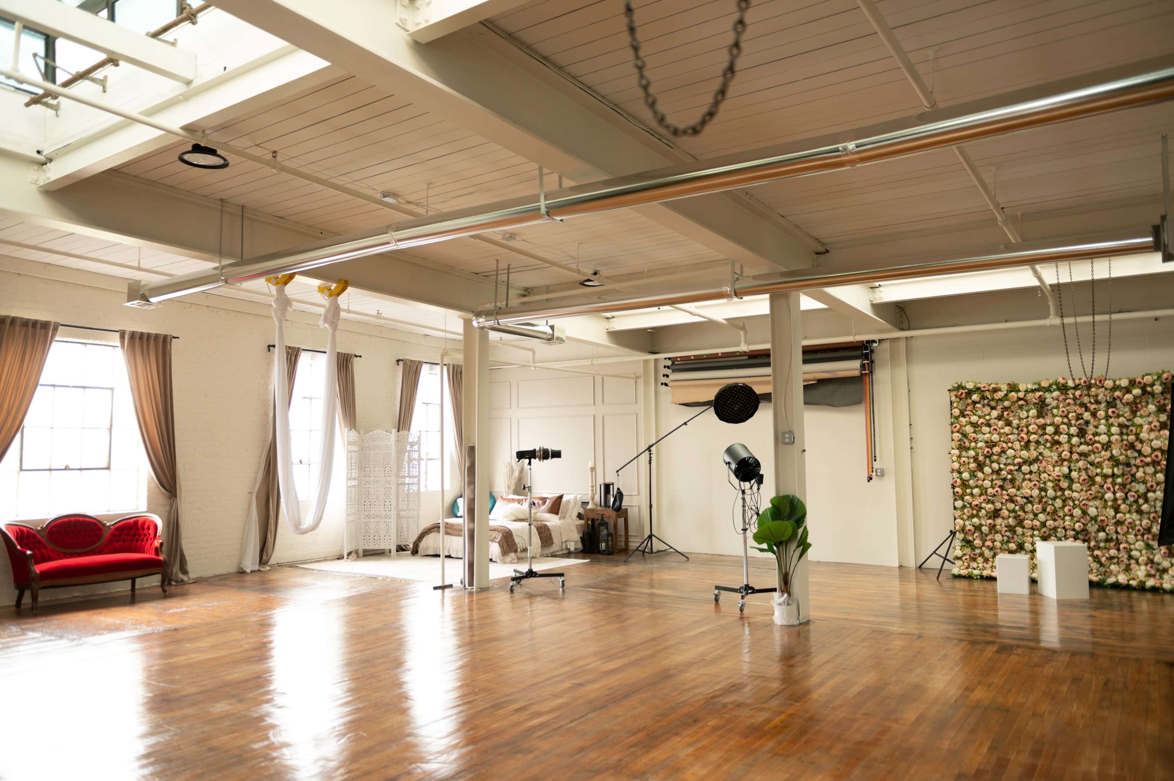A spacious studio with large windows, wooden floors, and a floral backdrop, along with a red couch and photography equipment.