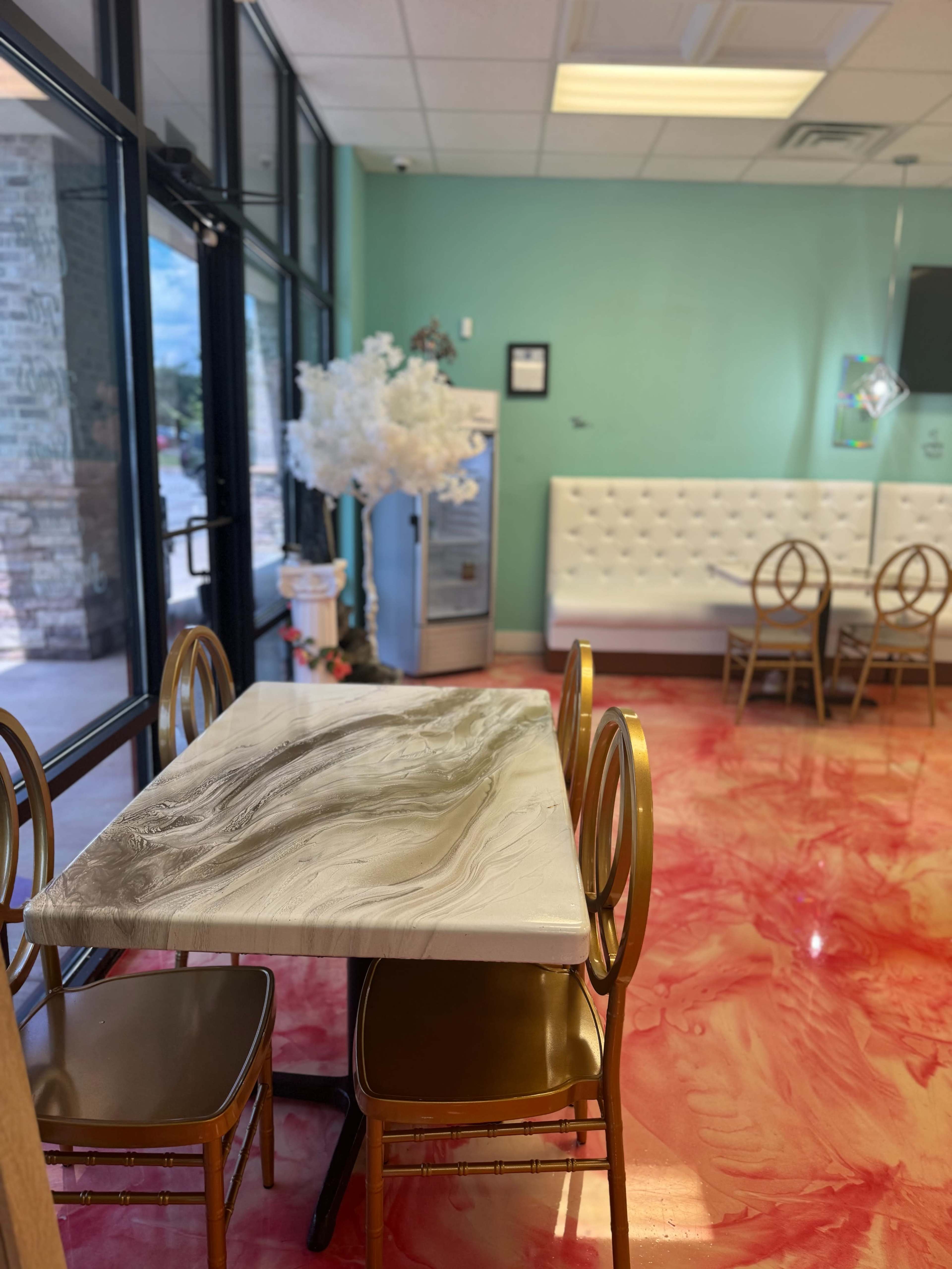 A brightly lit café interior with a glossy, marbled table, gold-framed chairs, and a pink, swirled floor.