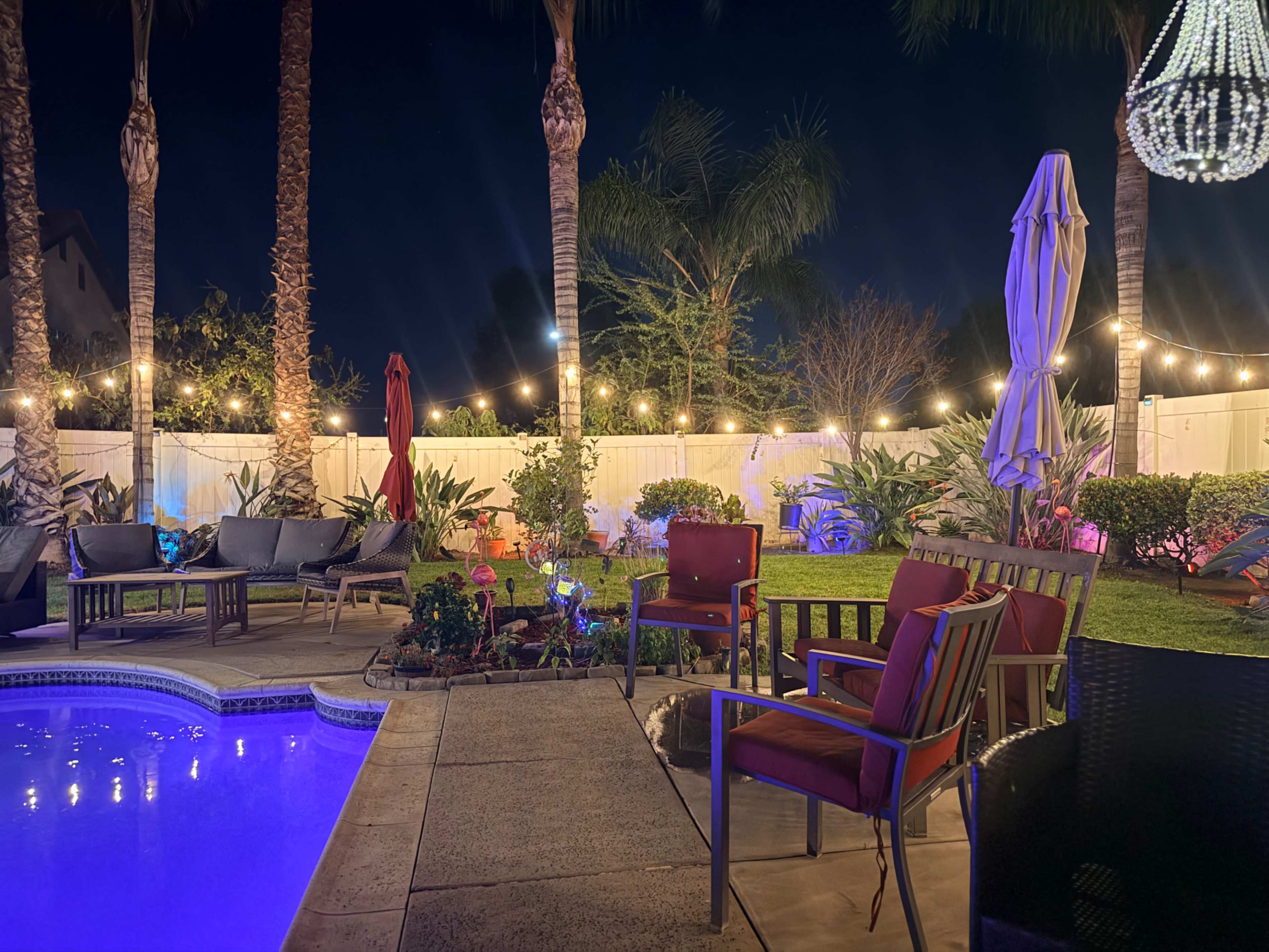 A backyard at night features a swimming pool with illuminated blue water, surrounded by patio furniture and decorative lights strung between palm trees.