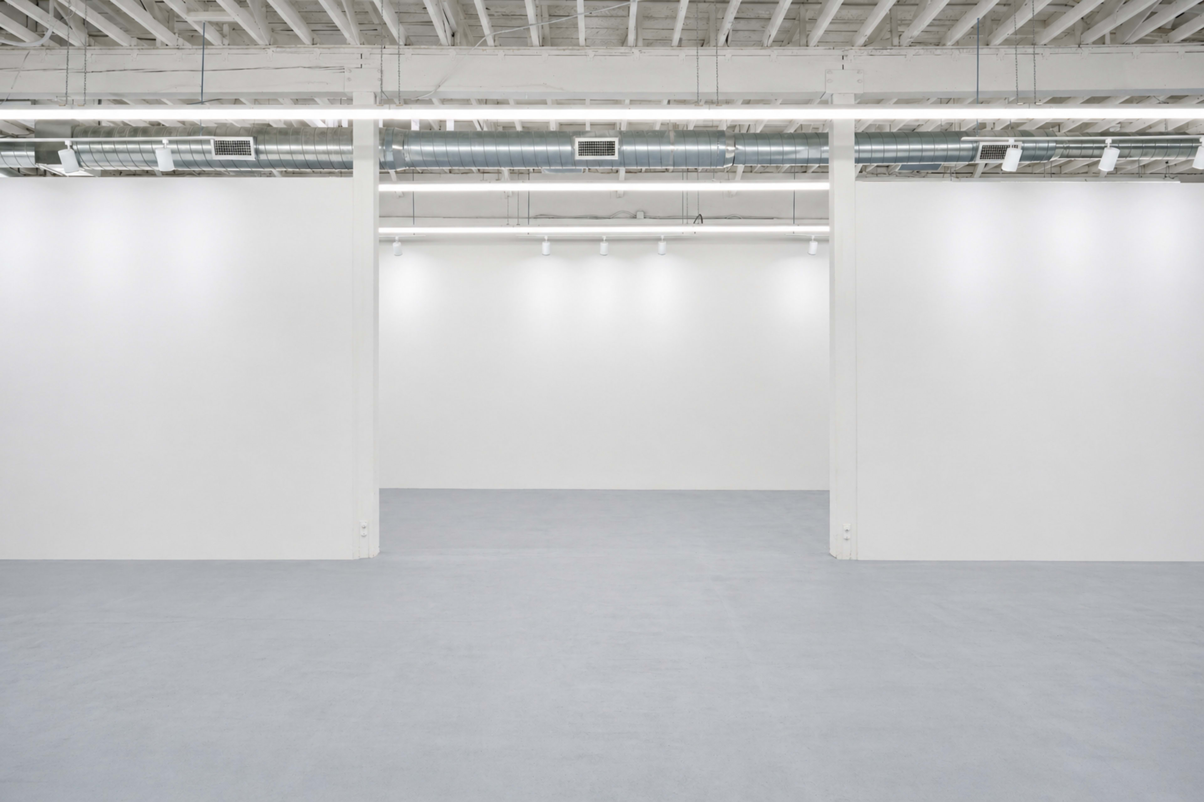 The image shows a spacious, empty gallery with white walls and a grey floor under bright overhead lighting.