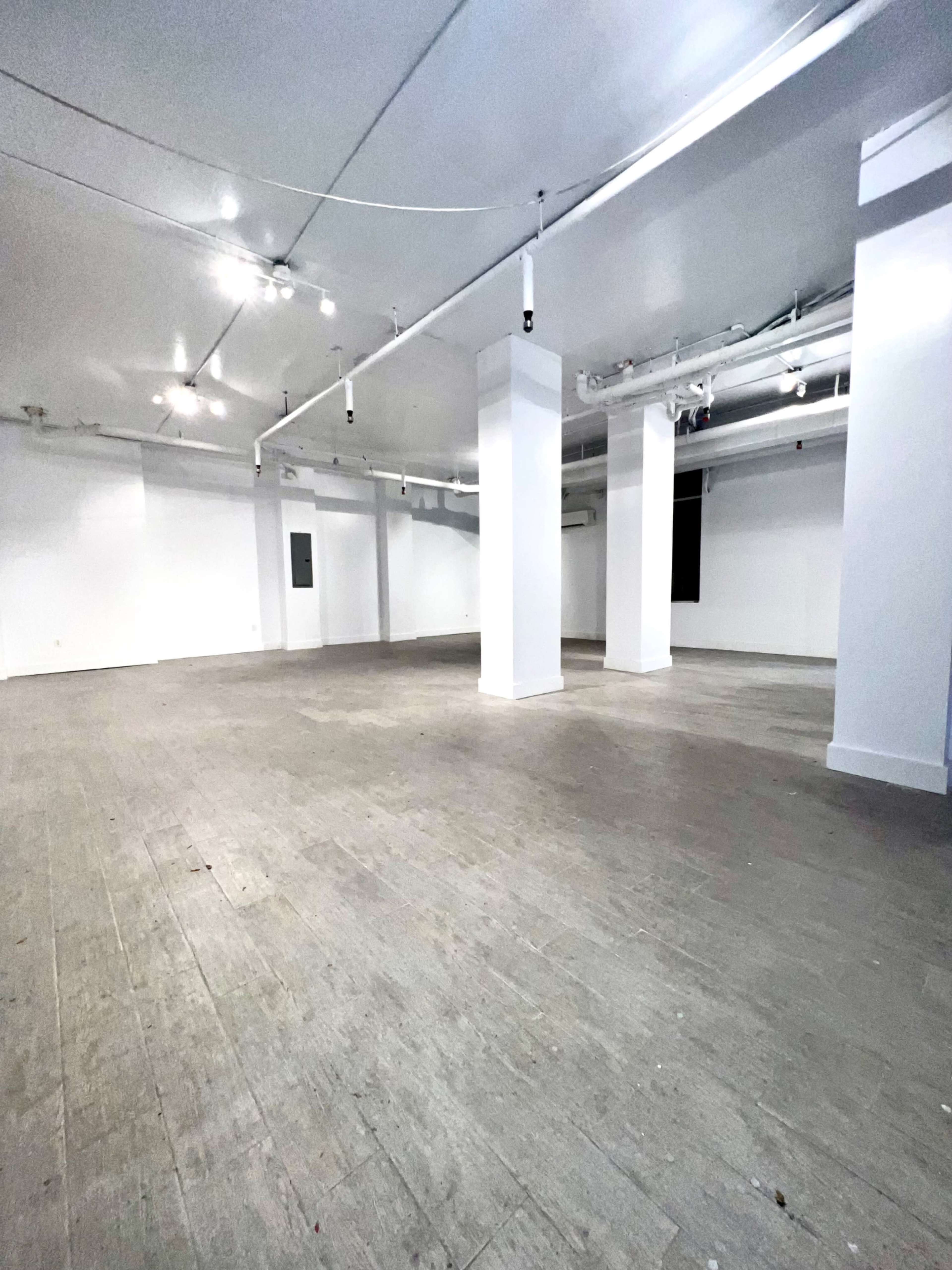 A large, empty commercial space with polished wooden floors and white walls, illuminated by overhead lights.