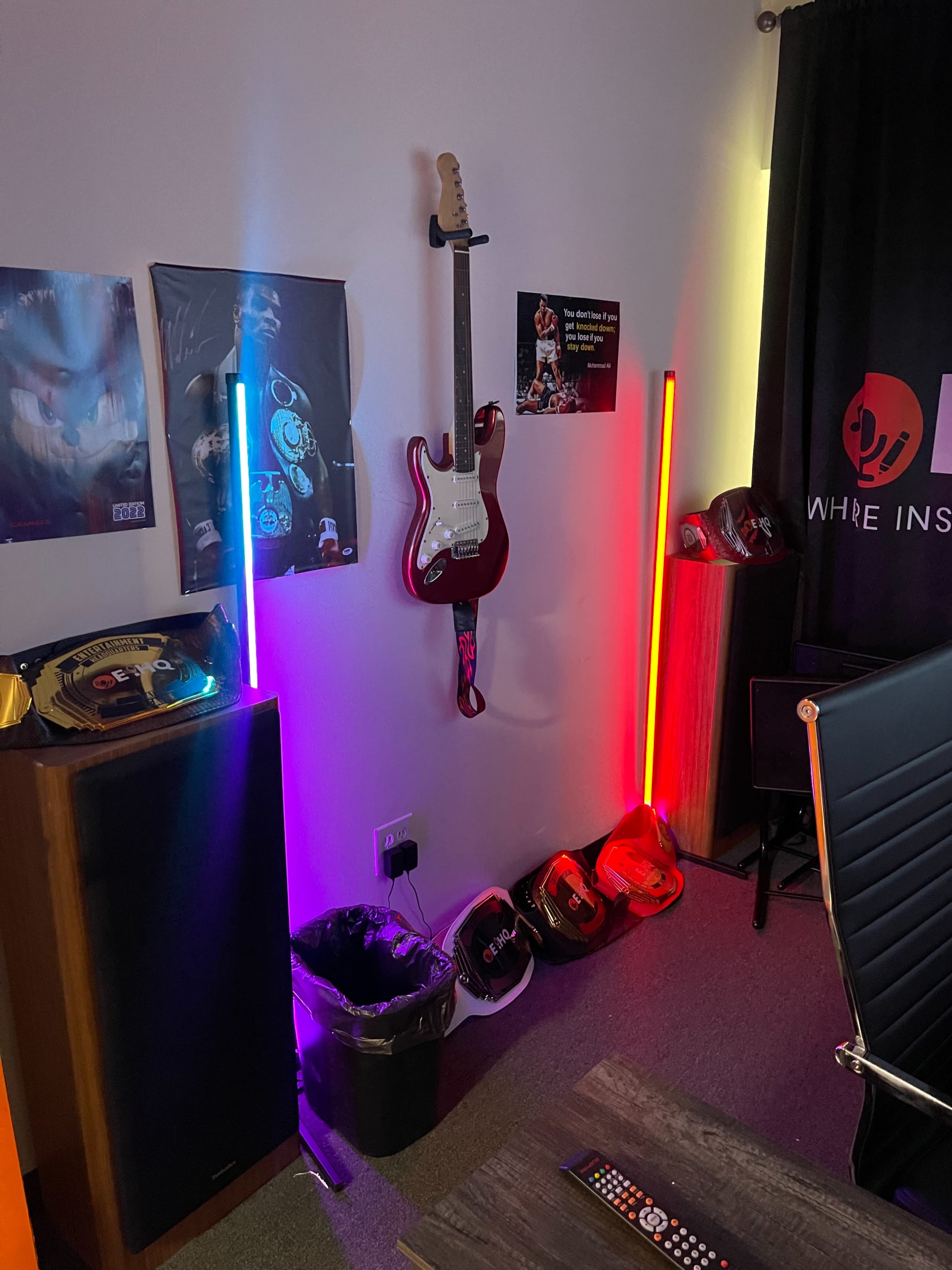 A corner of a room featuring an electric guitar mounted on the wall, colorful LED lights, a speaker, a trash bin, and various items on the floor.
