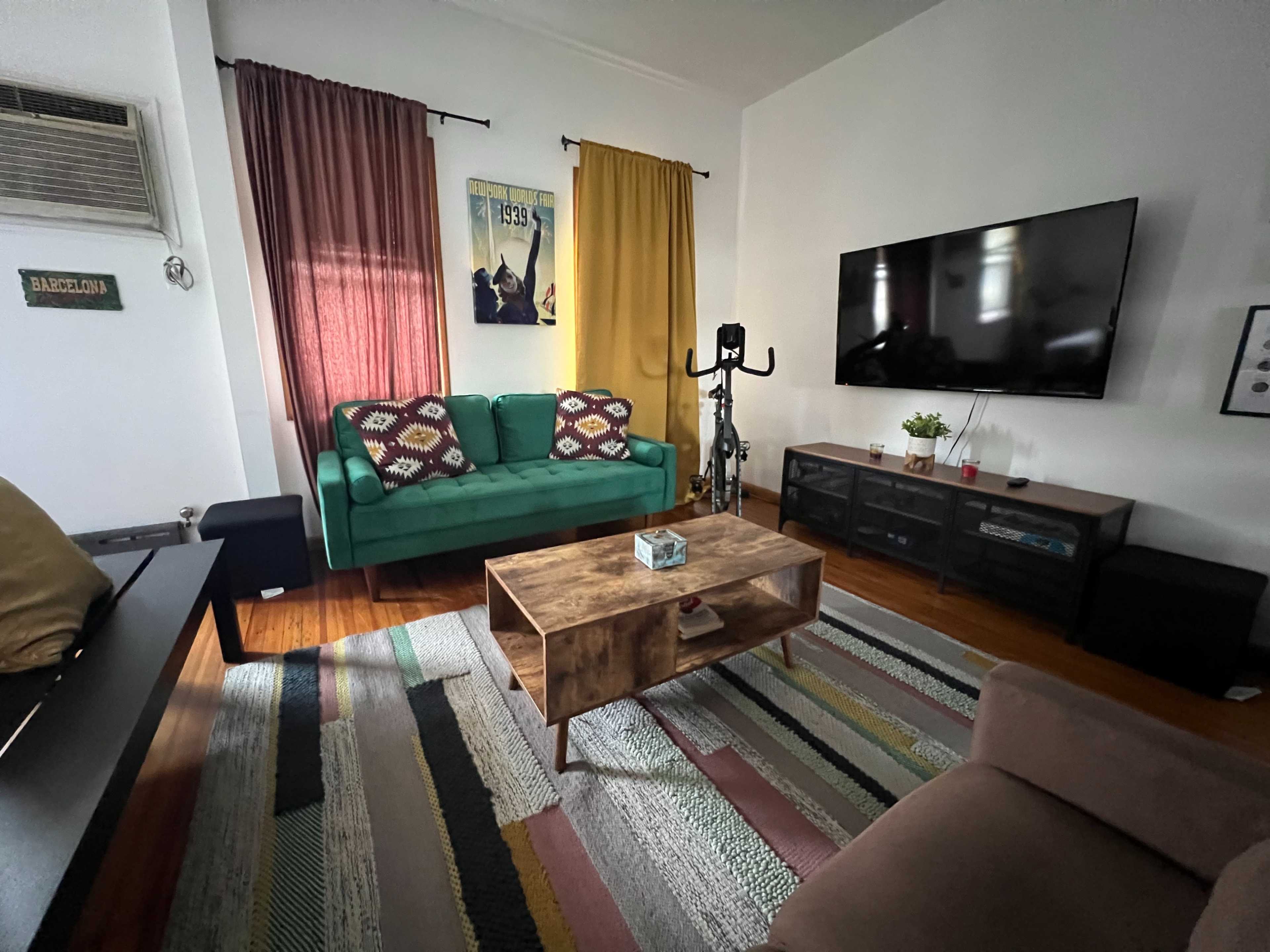 A living room features a green couch, a wooden coffee table, and a mounted television, with colorful curtains and a patterned rug on the floor.