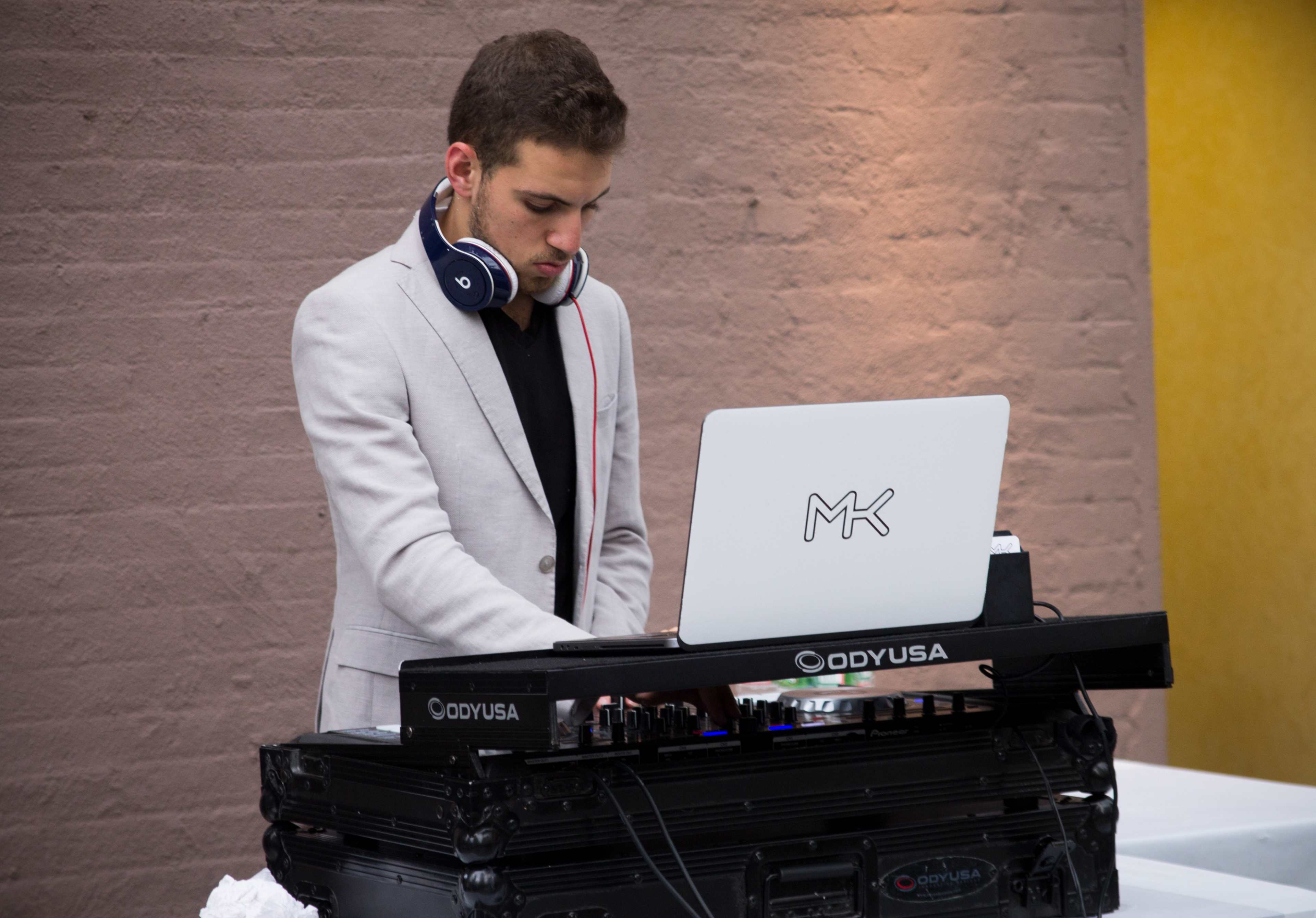 A DJ in a light-colored suit plays music on a turntable with a laptop in front of him, set against a textured wall.