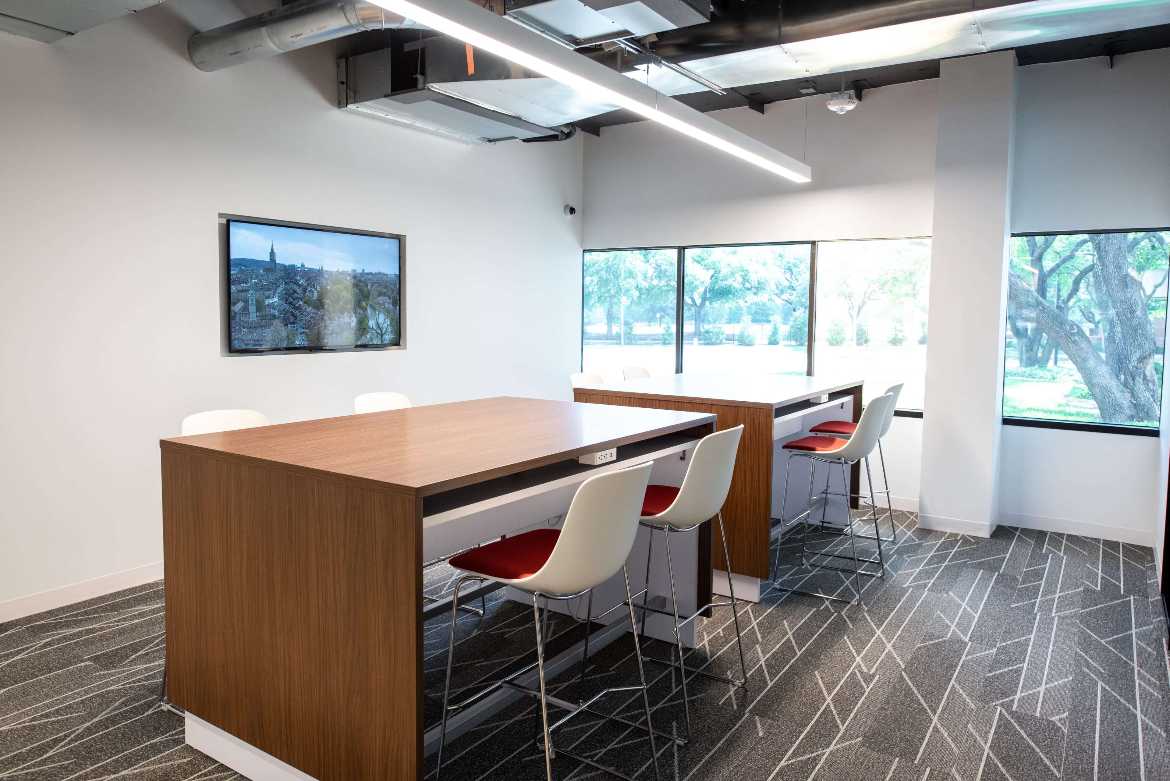 A modern office space with two wooden tables and white chairs, illuminated by overhead lighting, and large windows overlooking a green landscape.