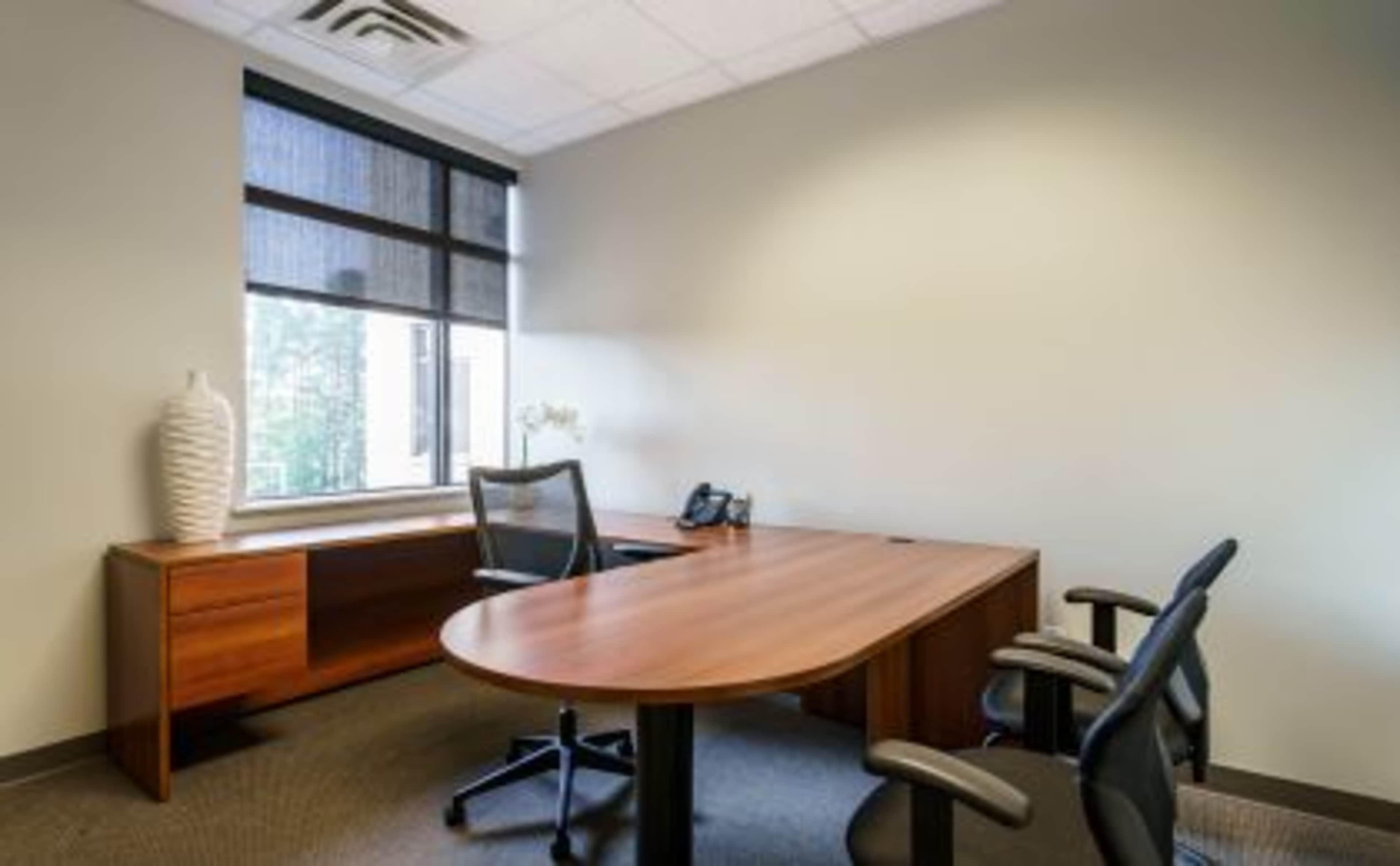 A minimalistic office space featuring a wooden desk, two black chairs, a window with blinds, and a decorative vase on a side table.
