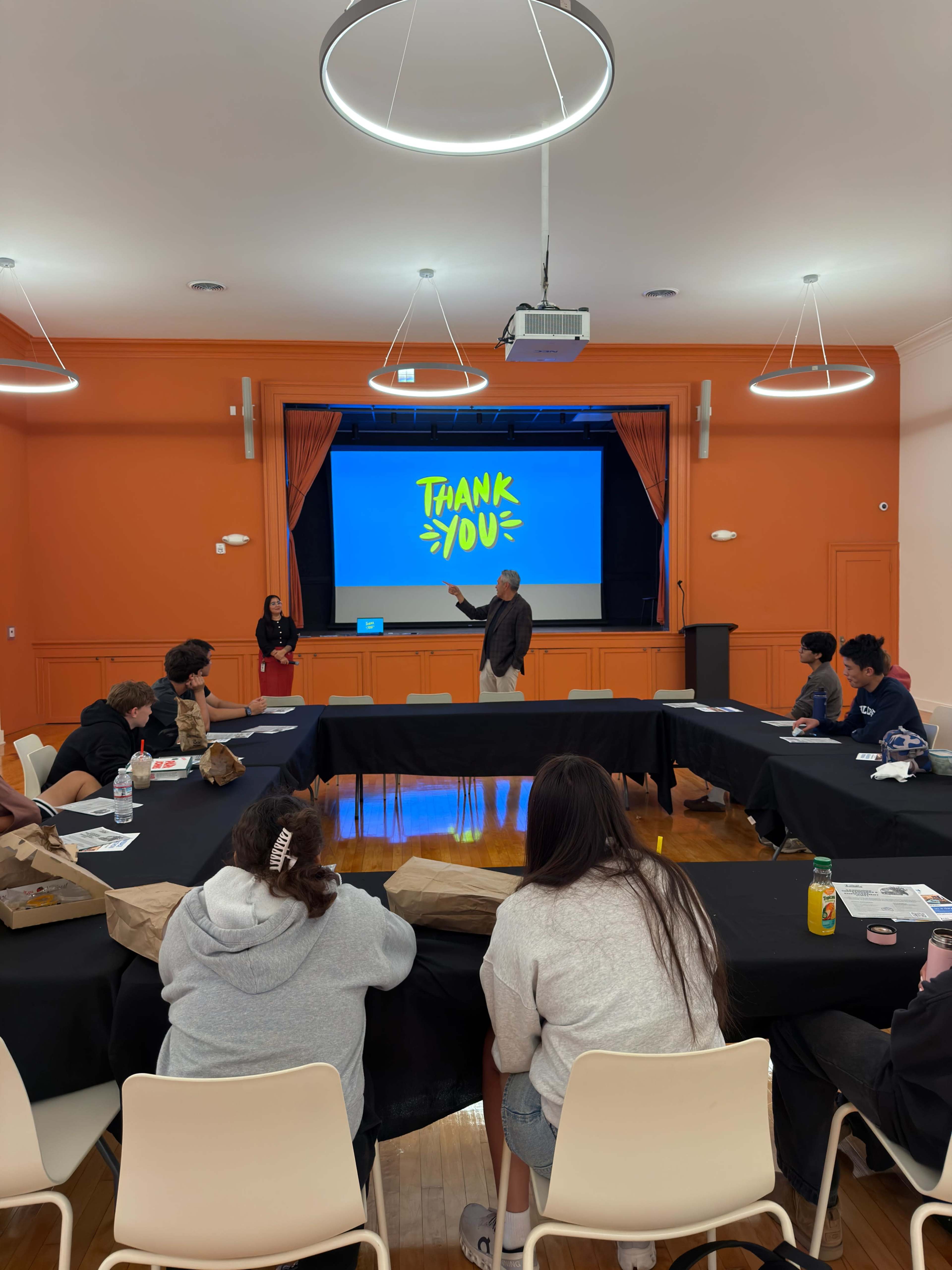 A speaker stands in front of a screen displaying "THANK YOU" while seated attendees listen in a classroom setting.