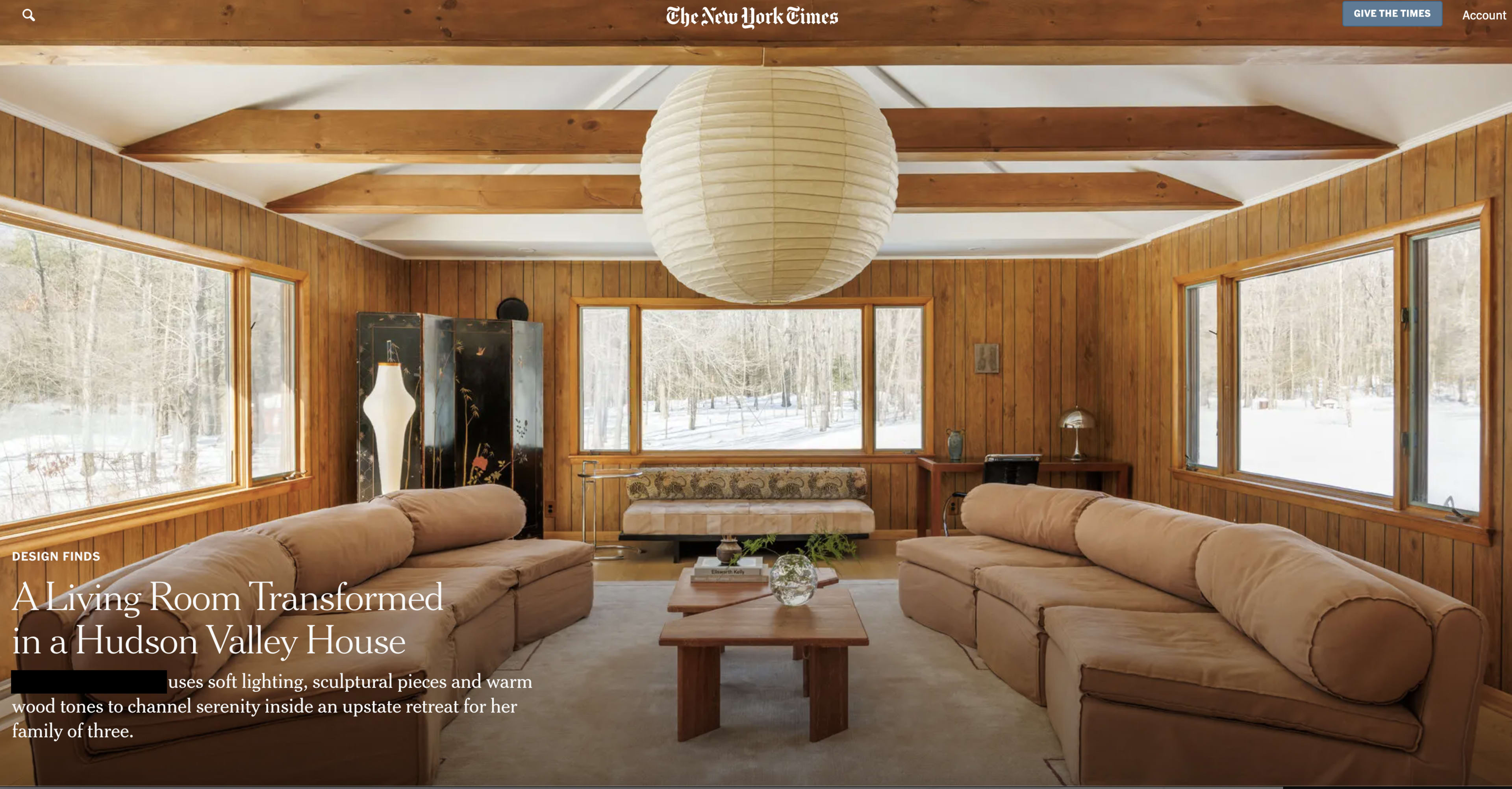 The image shows a spacious living room with wooden walls, large windows allowing natural light, and two beige sofas arranged around a central coffee table.