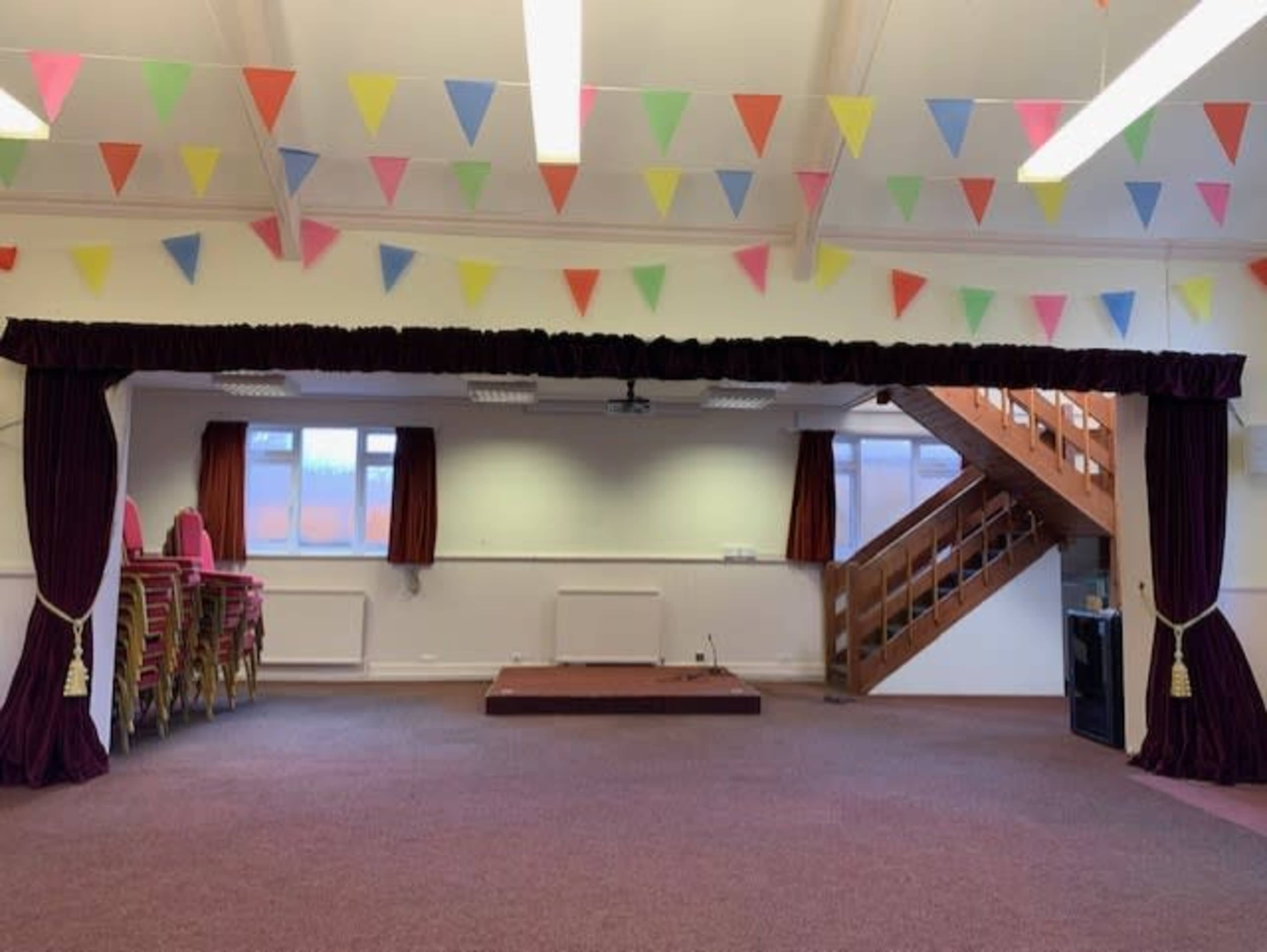 The interior of a hall features a stage framed by curtains, with colorful bunting hanging from the ceiling and chairs stacked against the wall.