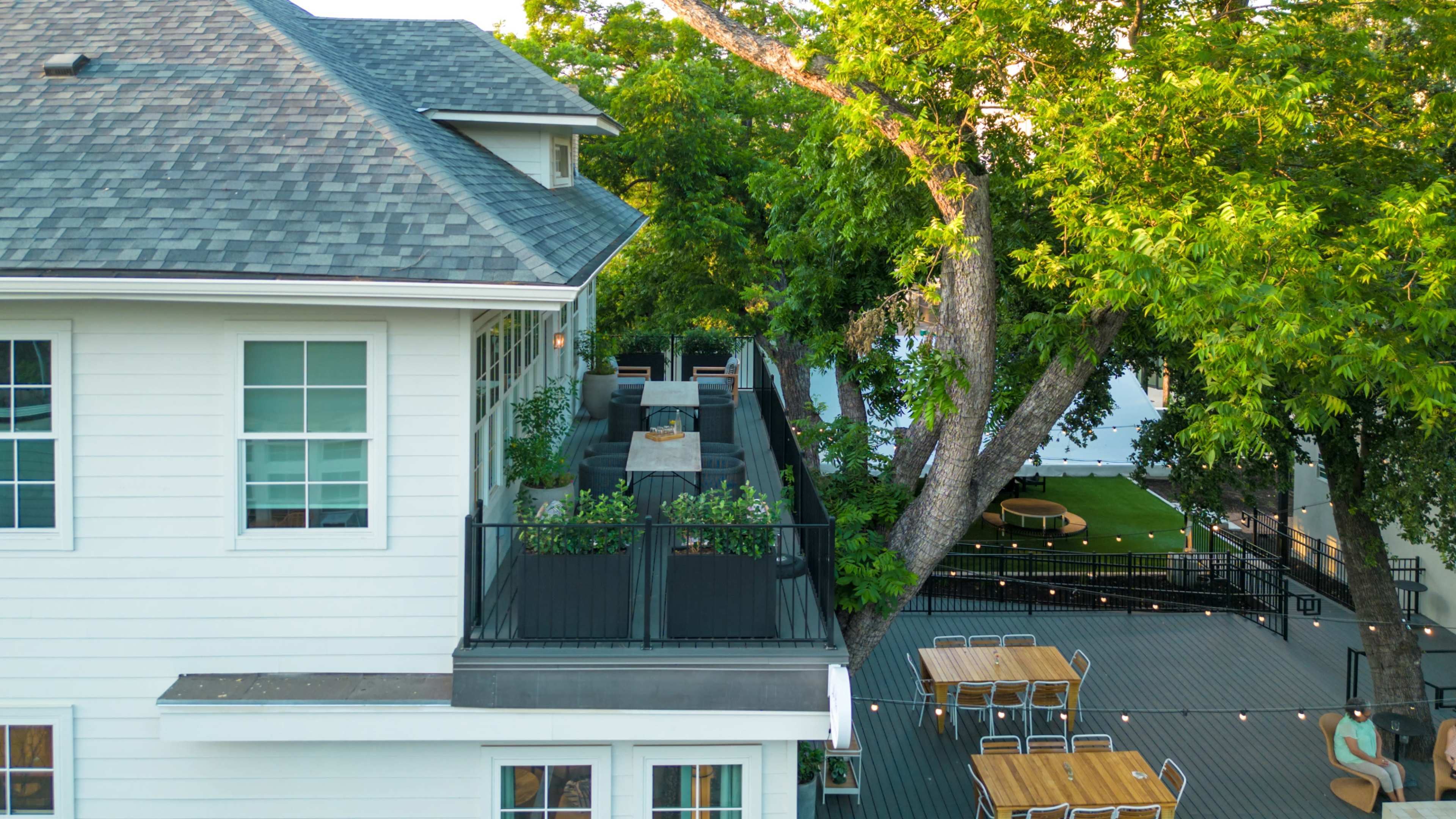 The image shows a multi-level house with a rooftop deck surrounded by trees, featuring outdoor seating and tables.
