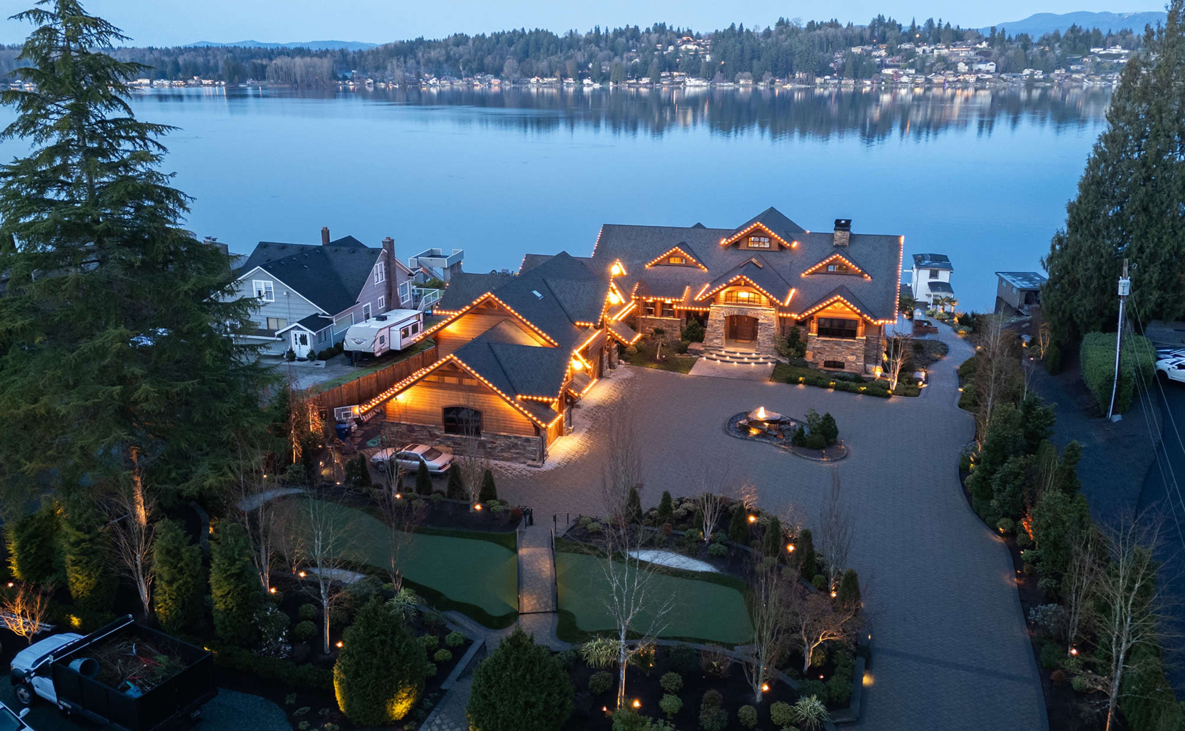 A large waterfront mansion with illuminated exterior features, surrounded by landscaped gardens and a paved driveway along a calm lake at dusk.
