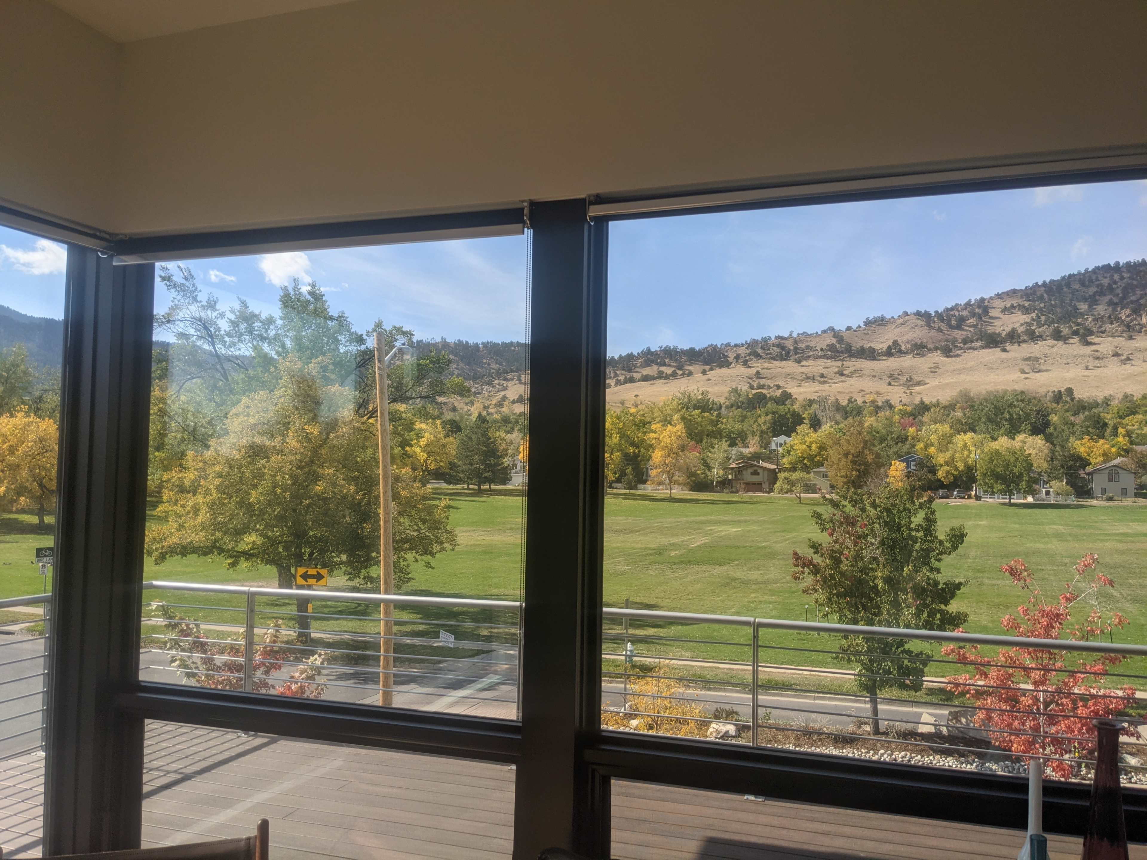 A view through large windows overlooking a grassy field surrounded by trees with autumn foliage and distant hills.