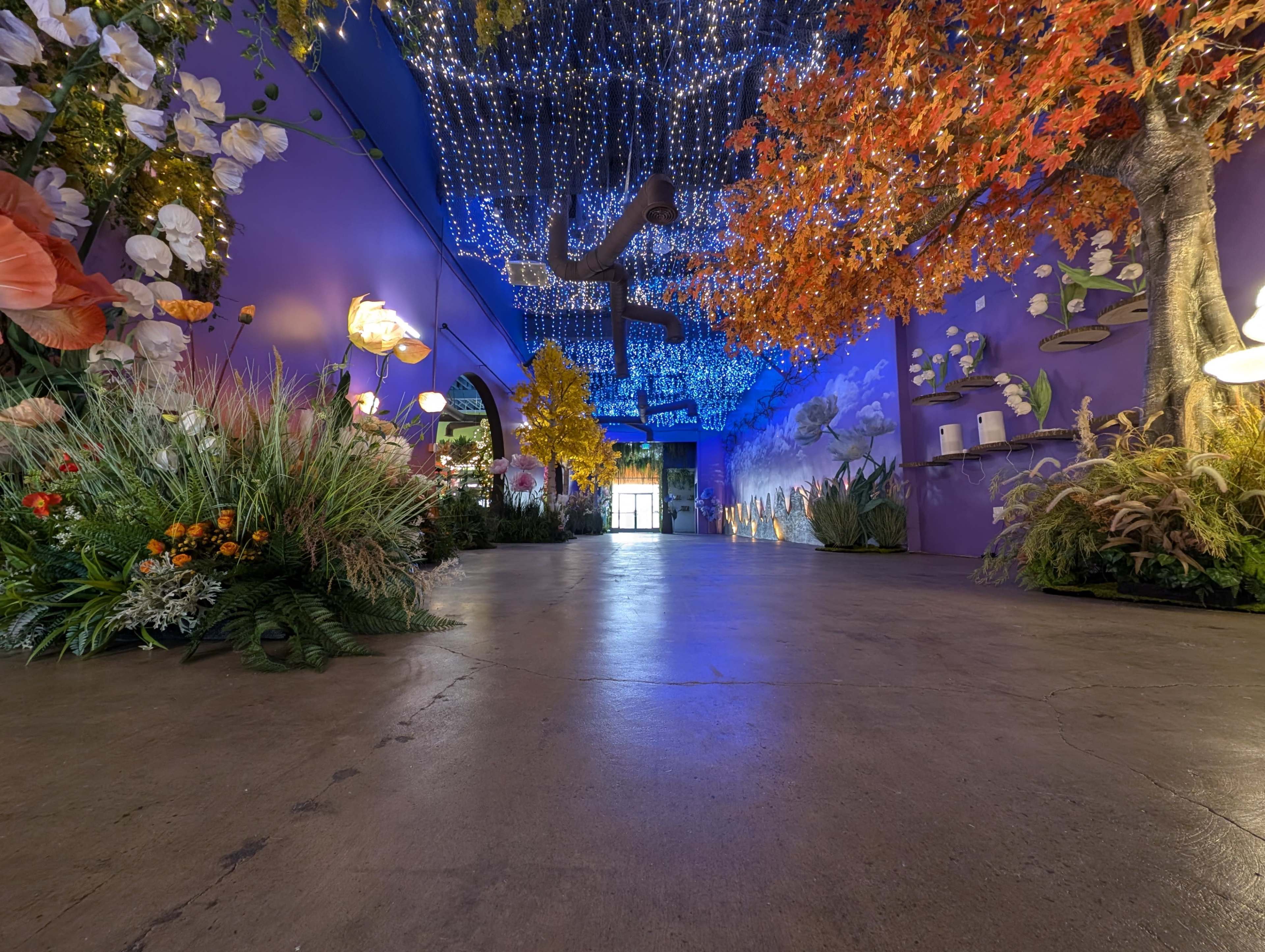 The image shows a vibrant, colorful hallway adorned with artificial flowers, plants, and decorative lighting, leading toward an exit at the far end.