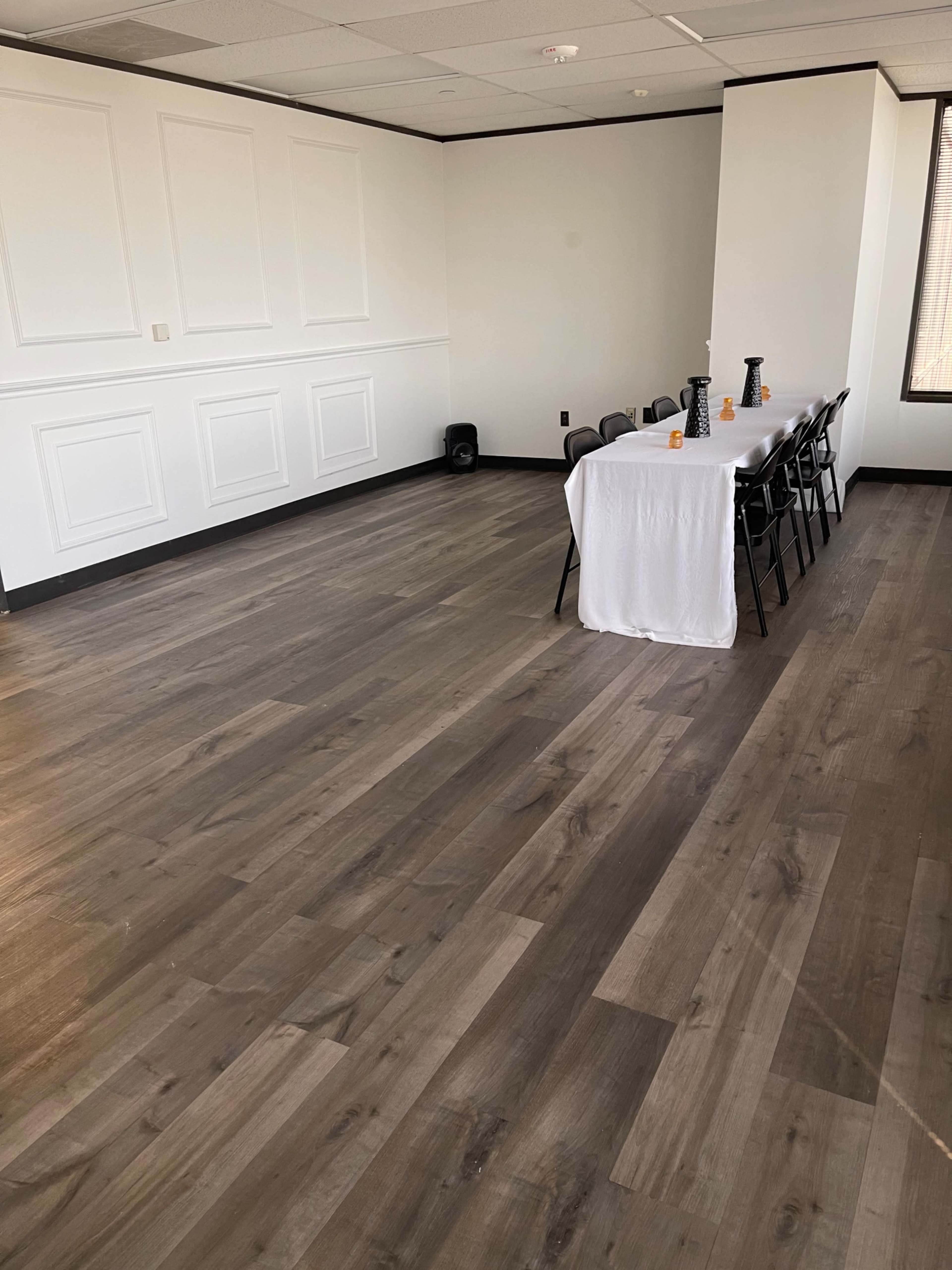 The image shows a spacious, empty room with gray wooden flooring and a long table set with a white tablecloth and black chairs.
