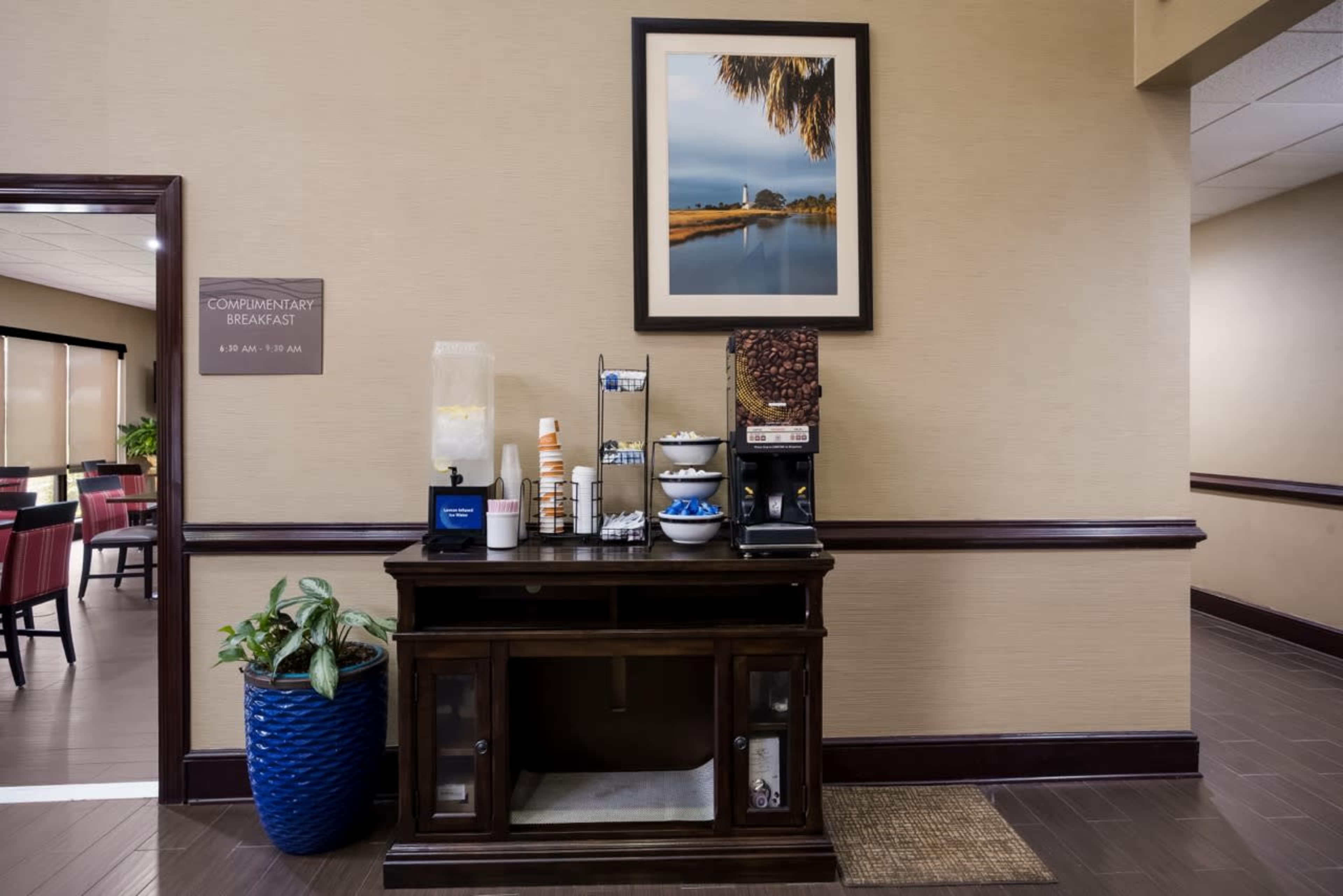 The image shows a breakfast area in a hotel with a wooden console displaying coffee, condiments, and bowls, alongside a water dispenser and a wall-mounted photo.