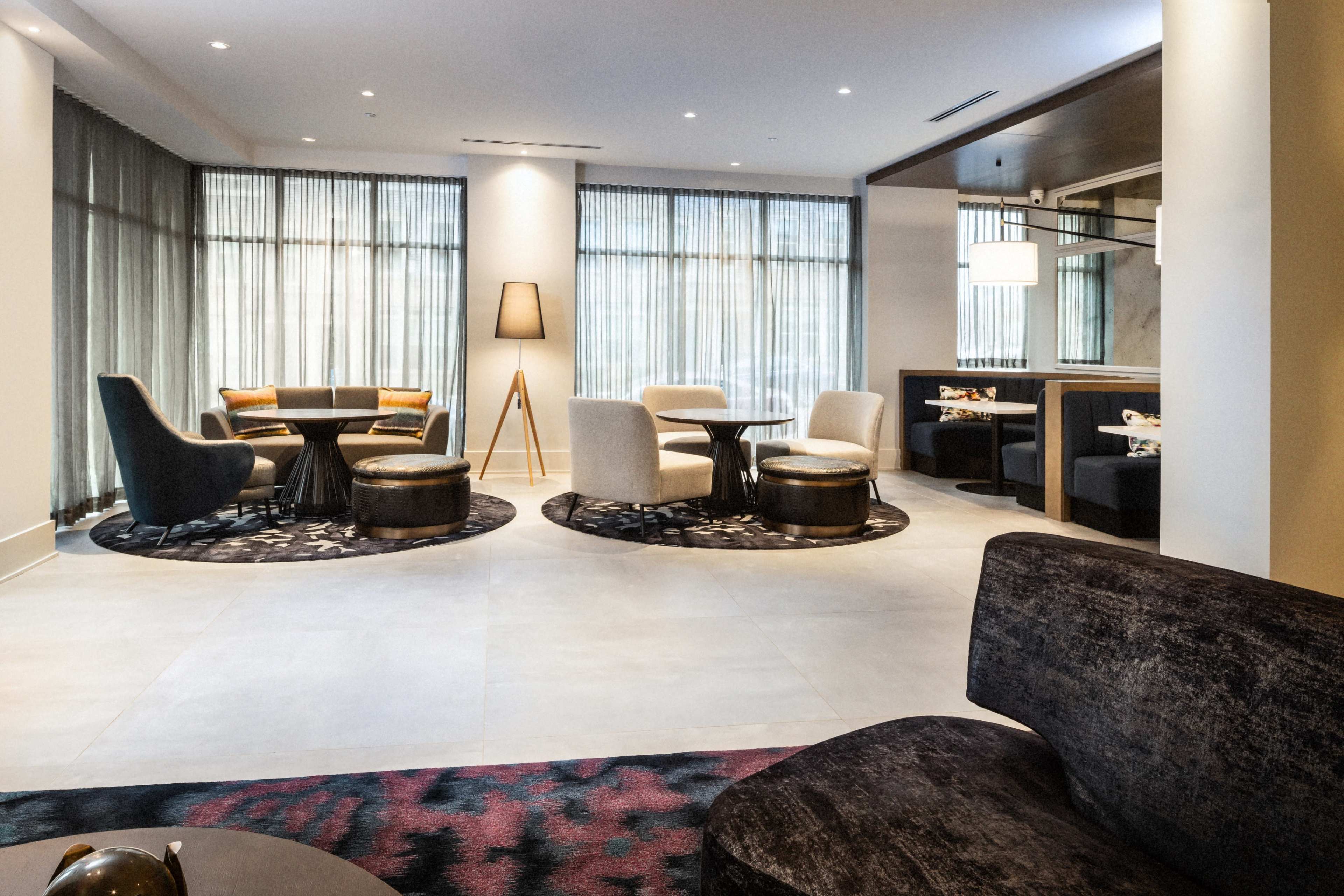 A contemporary lounge area featuring various seating arrangements, including chairs and tables, set against large windows with sheer curtains.